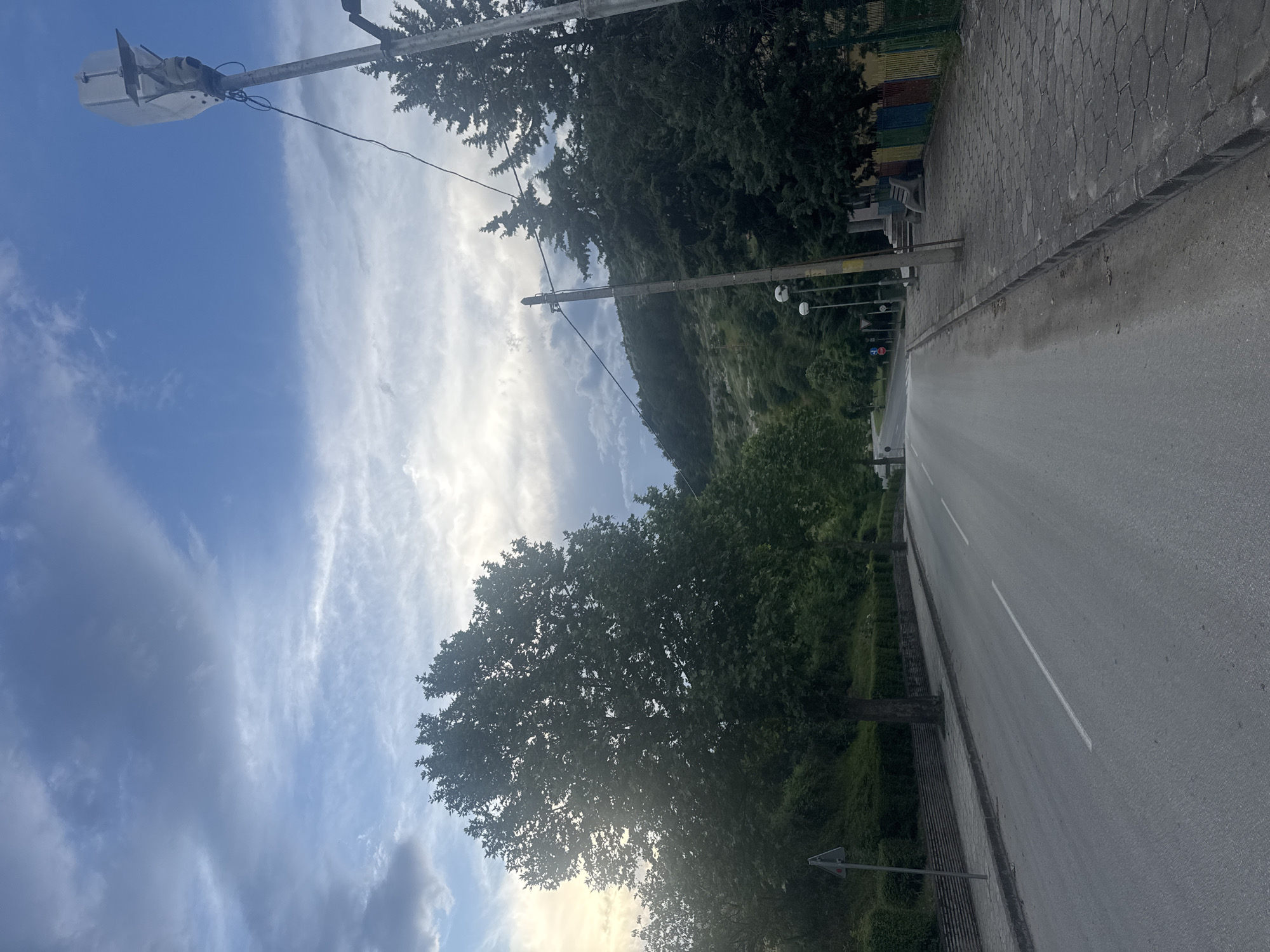 The image shows a street in Ivaylovgrad, Bulgaria, lined with trees and vegetation. A gray paved sidewalk runs along the right side of the street, alongside a gray asphalt road with a single white dividing line. A mountain slope covered in trees is visible in the background. Power lines and a street light are visible on the left side of the frame against a cloudy blue sky.