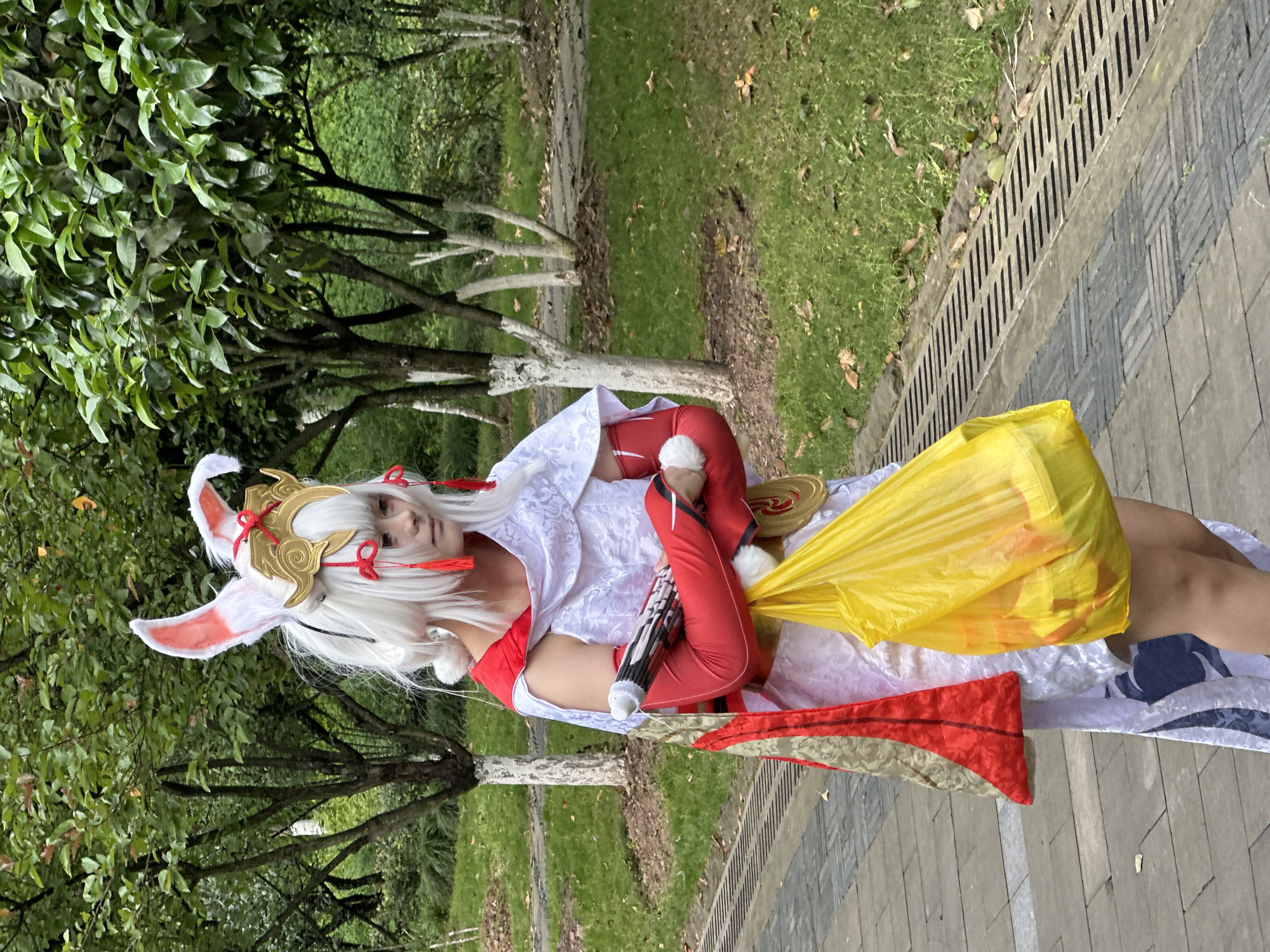 The image shows a person in cosplay, standing on a paved pathway in what appears to be a park or garden. The background includes greenery, trees, and a grassy area. The pathway is made of grey paving stones. The person is wearing a white wig with rabbit ears, a golden headpiece, and red and white attire. They have a yellow bag or piece of fabric draped over their shoulder. The image was taken outdoors, possibly in Liangjiang, China, given the user's prompt.