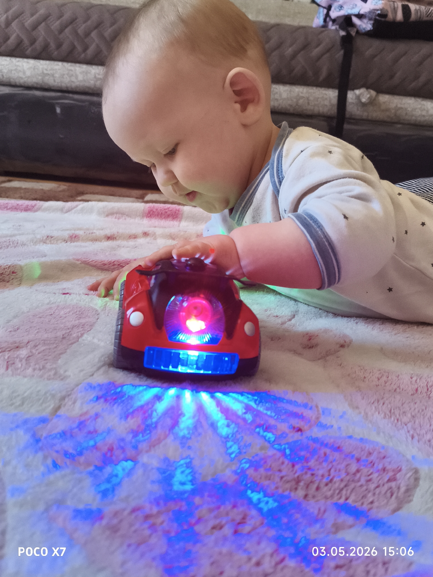 A baby lies prone on a pink and white blanket indoors, with a red and black toy car illuminating the blanket with blue and red light. The baby's left hand rests on the car. A piece of furniture is visible in the background. The image includes the text "POCO X7" and "03.05.2026 15:06." There is no contextual information in the image that allows me to determine the location as Chaikovsky, Russia.