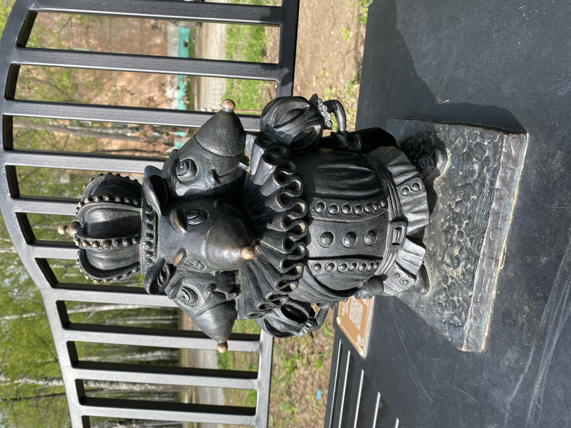 The image shows a bronze sculpture of a character from a Russian fairy tale located in Chaikovsky, Russia. The sculpture depicts a rat dressed in royal attire, including a crown and ruffled collar. The rat is holding a small object and standing on a textured base. The sculpture is positioned on a black surface next to a metal railing. The background includes blurred trees and buildings.