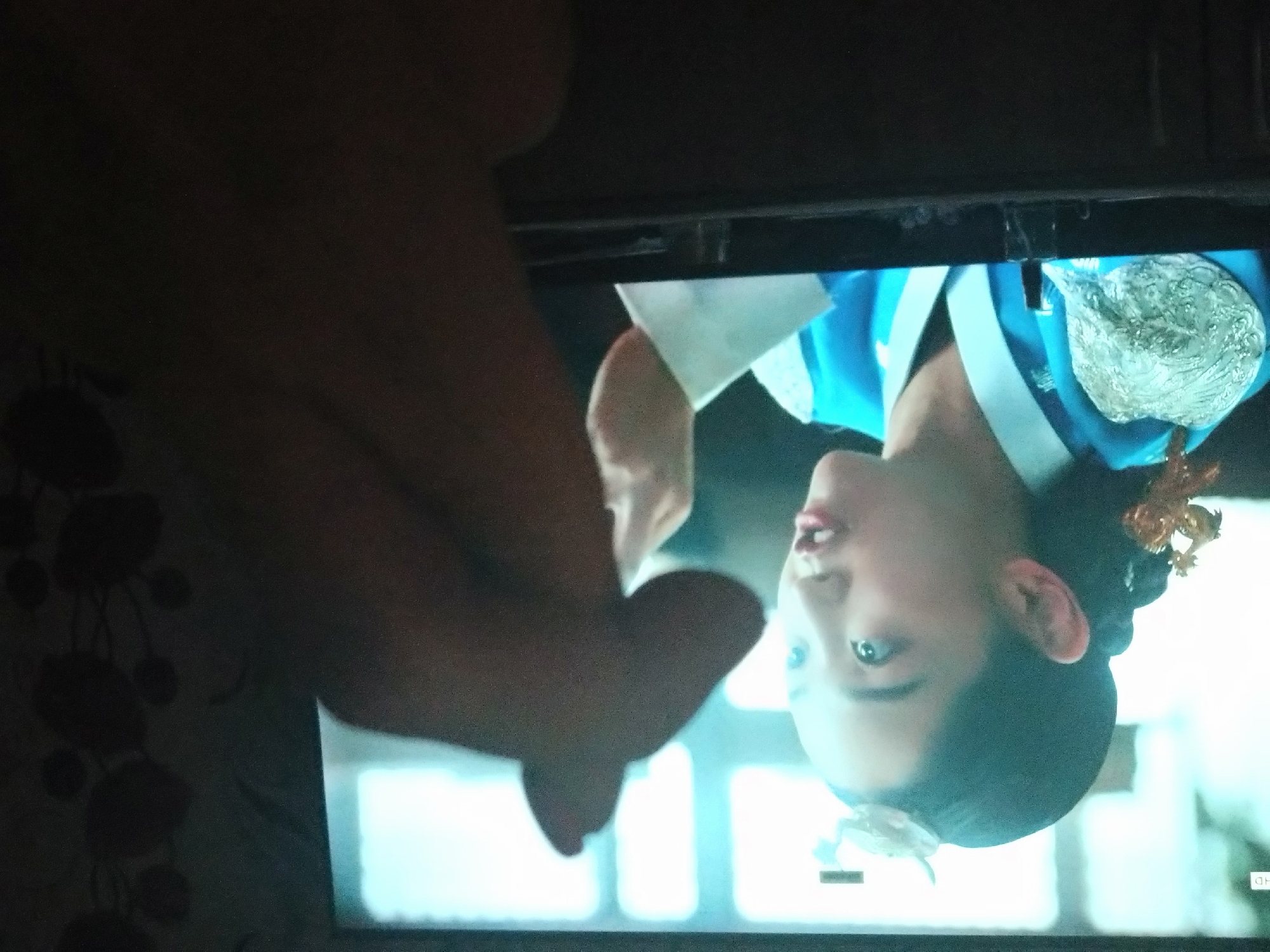 A person's hand obscures part of a TV screen that shows the face of a woman in a traditional Korean dress. The woman's face is oriented upside down in the image. The setting or specific location is not discernible from the image.