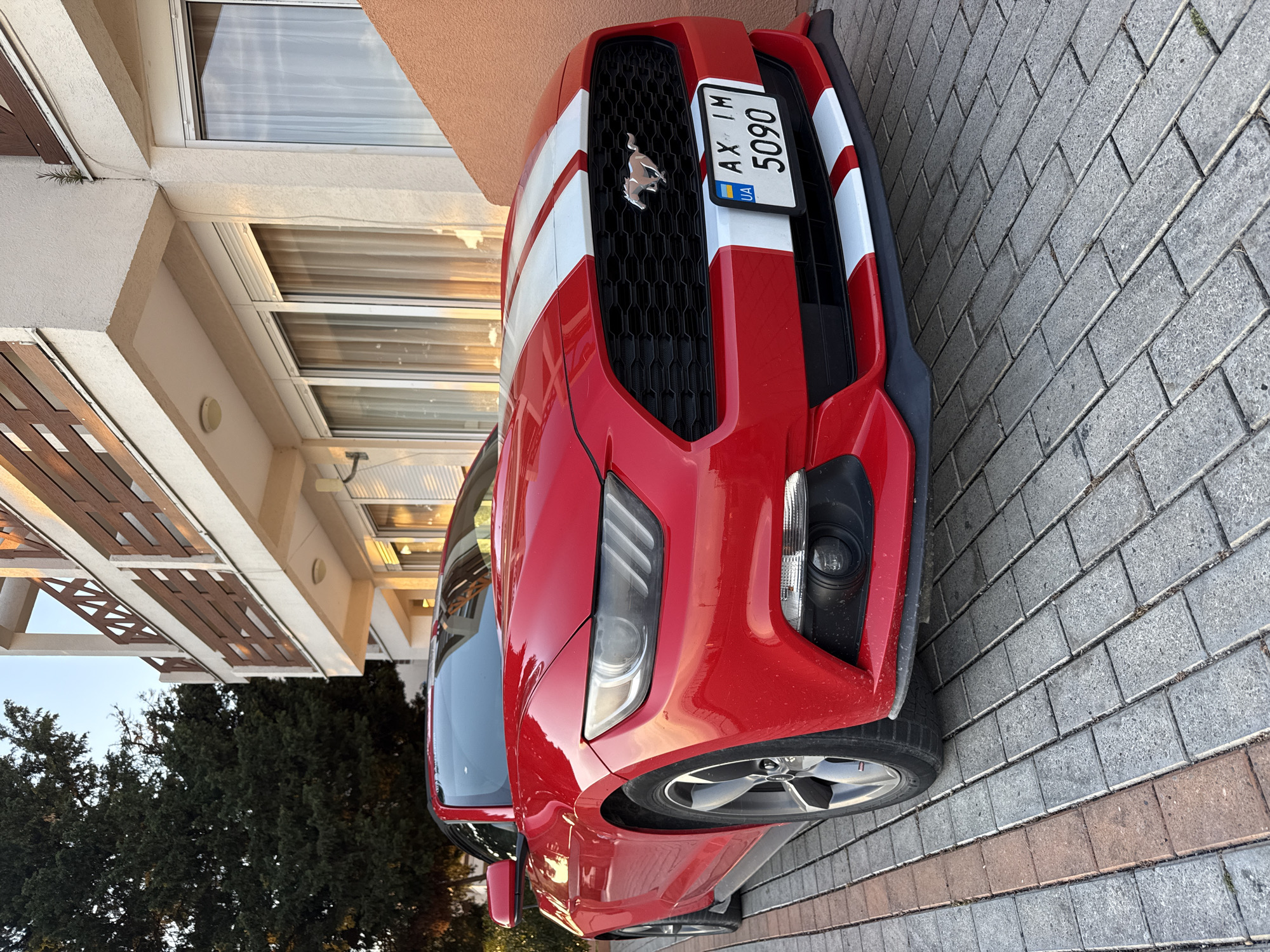 A red Ford Mustang with white stripes is parked on a gray brick driveway in front of a modern building. The car's license plate reads "AX IM 5090" with the "UA" designation, indicating it's a Ukrainian vehicle. The building has multiple floors with windows and a tan facade. Trees are visible in the background on the left side of the frame. The image orientation suggests the car is parked at an angle relative to the building. The location is not indicated in the image. The city of Šušanj, Montenegro, cannot be verified based on visual elements in the image.