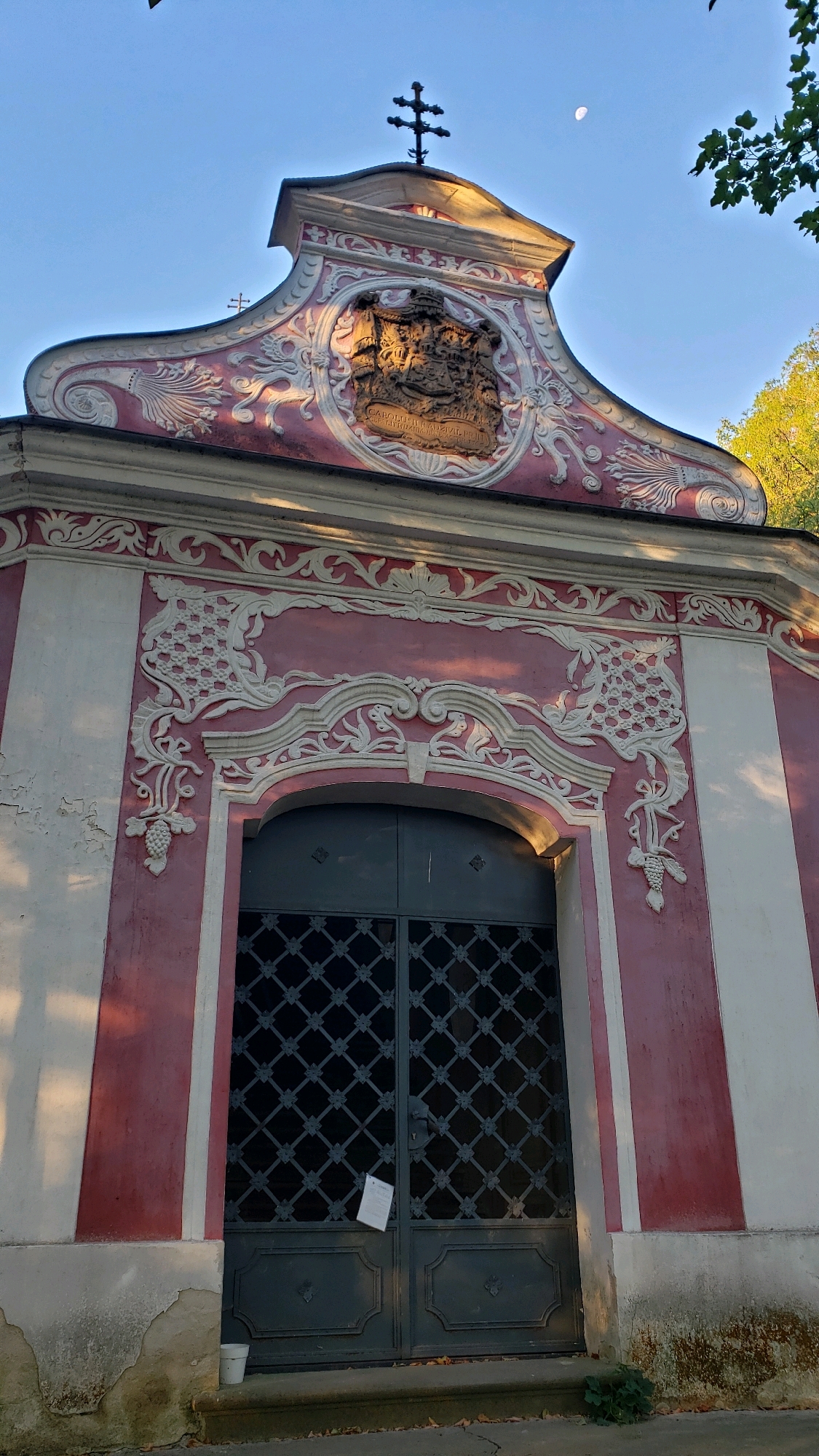 The image shows the exterior of a building with pink and white walls, decorated with ornate stucco work. A large, dark metal gate with a diamond pattern is visible in the center of the facade. Above the gate, the stucco work features intricate scrollwork and floral motifs, with a prominent coat of arms in a circular frame at the top. A double-barred cross stands on the apex of the roof. The building appears to be a chapel or a mausoleum. The scene is set against a clear blue sky with a sliver of the moon visible. Trees and foliage are present in the background, suggesting an outdoor setting. The image is taken from a low angle, looking up at the building. The architectural style suggests historical significance, possibly Baroque or Rococo. The condition of the lower part of the facade shows some signs of wear and tear. The building is located in the Catholic Cemetery in Prešov, Slovakia.