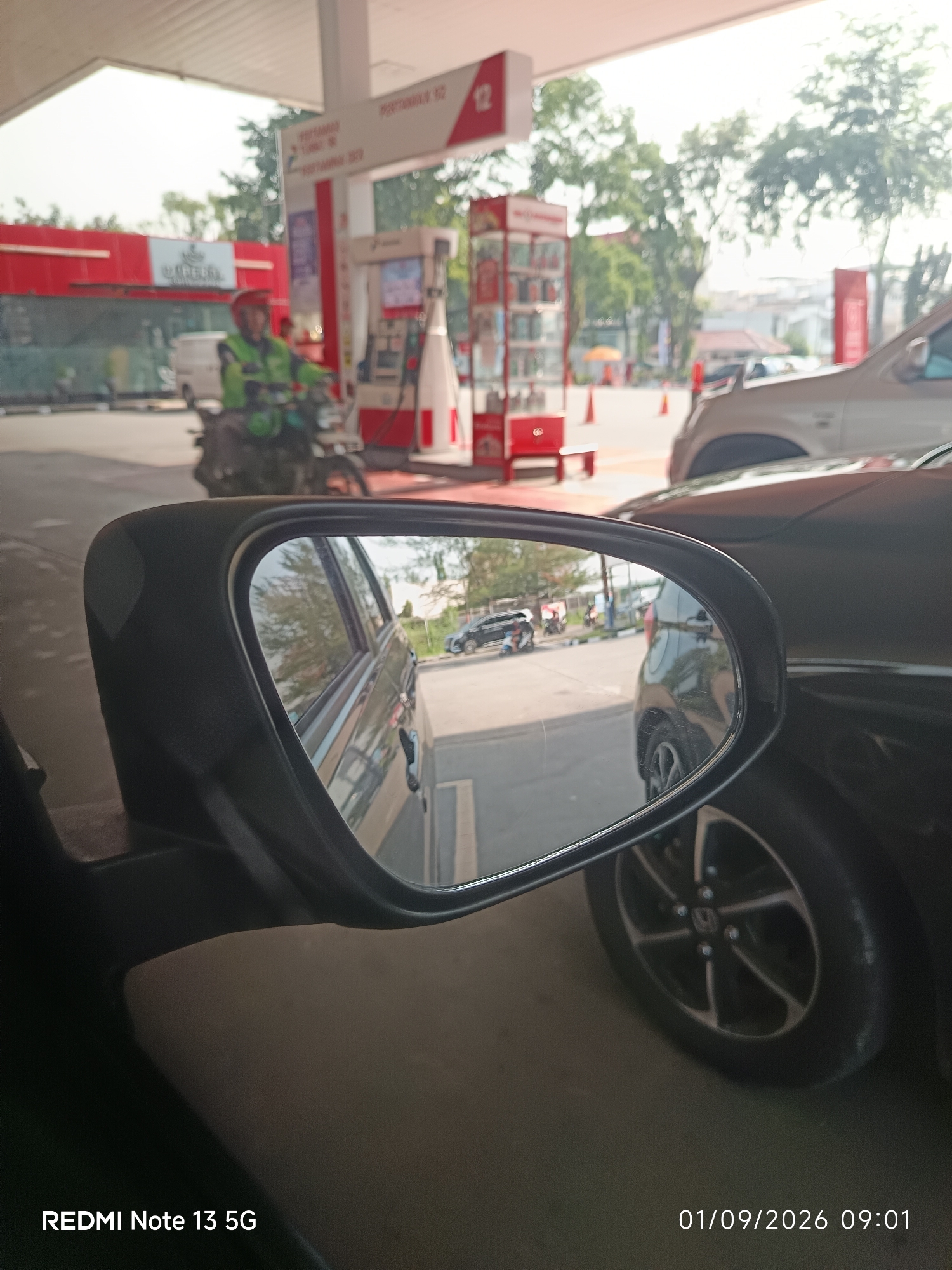 The image depicts a gas station in the city of Medan, Indonesia, viewed from the perspective of a car. In the foreground, a side-view mirror reflects a black car and the surroundings, including a gas pump and other vehicles. A person wearing a green vest and a red helmet is seen riding a motorcycle in the background, near the gas pumps. The gas station appears to be open, with several vehicles present and signs indicating it is a Pertamina station. The overall scene is one of everyday activity at a public transportation hub in a bustling urban environment.