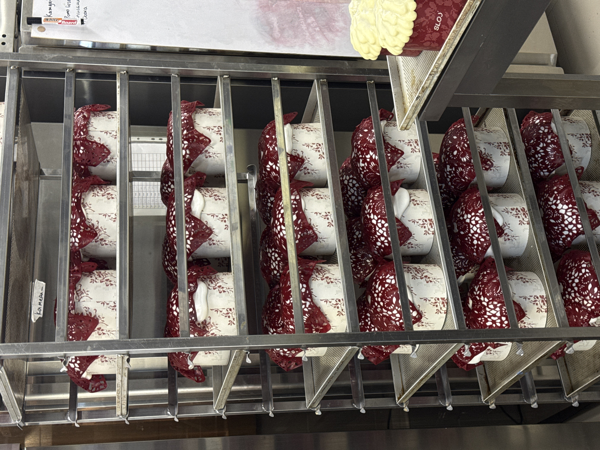 The image shows rows of neatly arranged containers, likely food or drink containers, inside a stainless steel racking system. The containers appear to be white with a patterned design, and many have a red, lace-like adornment around them. The scene is indoors. The upper-left corner features a whiteboard with handwritten notes in a language other than English. The word "SLOJ" is visible on one of the containers in the upper-right area of the photo. A small label is seen with the word "Kamera" on it. The image location is Belgrade, Serbia.