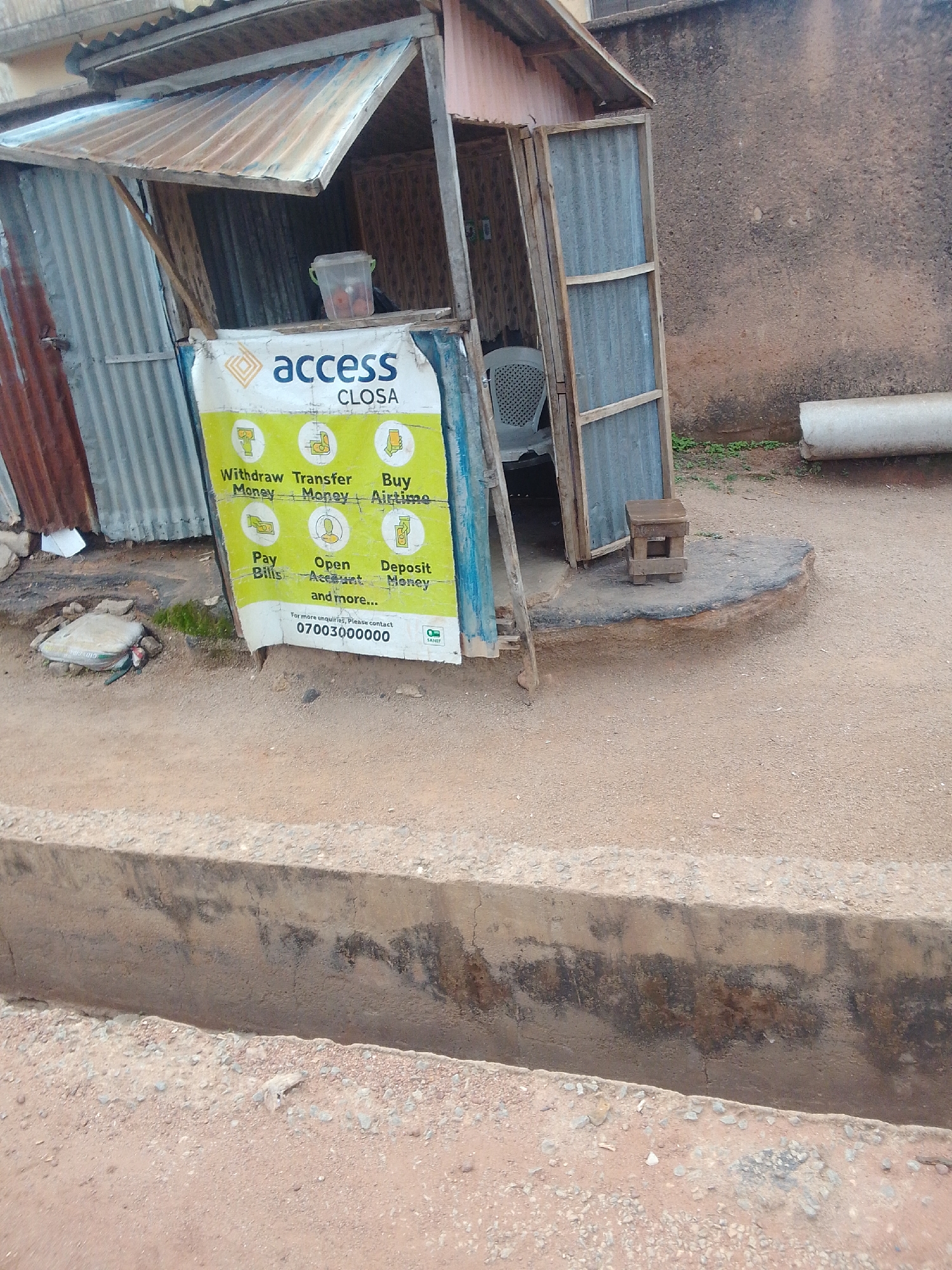 This image displays an outdoor scene in the city of Jos, Nigeria, featuring a small, open-fronted kiosk. The structure is constructed from corrugated metal sheets and wooden framing, with a corrugated metal awning extending from the roof over the front. Inside the kiosk, a white plastic chair is visible, along with a clear plastic container resting on a counter. A white and yellow banner, branded "access CLOSA," is affixed to the front of the kiosk, advertising services such as "Withdraw Money," "Transfer Money," "Buy Airtime," "Pay Bills," "Open Account," and "Deposit Money," along with the contact number "07003000000" and "SAJEF" branding.
A small, square wooden stool is positioned on a raised, dark-rimmed concrete slab directly in front of the kiosk. The ground immediately surrounding the structure is unpaved, composed of reddish-brown soil and scattered gravel. Sparse green vegetation is present in isolated patches on the ground. To the right of the kiosk, a rough-textured, light brown wall is visible, and a segment of a concrete pipe rests on the ground adjacent to the wall. In the foreground, a concrete drainage ditch, exhibiting dark staining and minor damage, traverses the scene. No individuals are present.