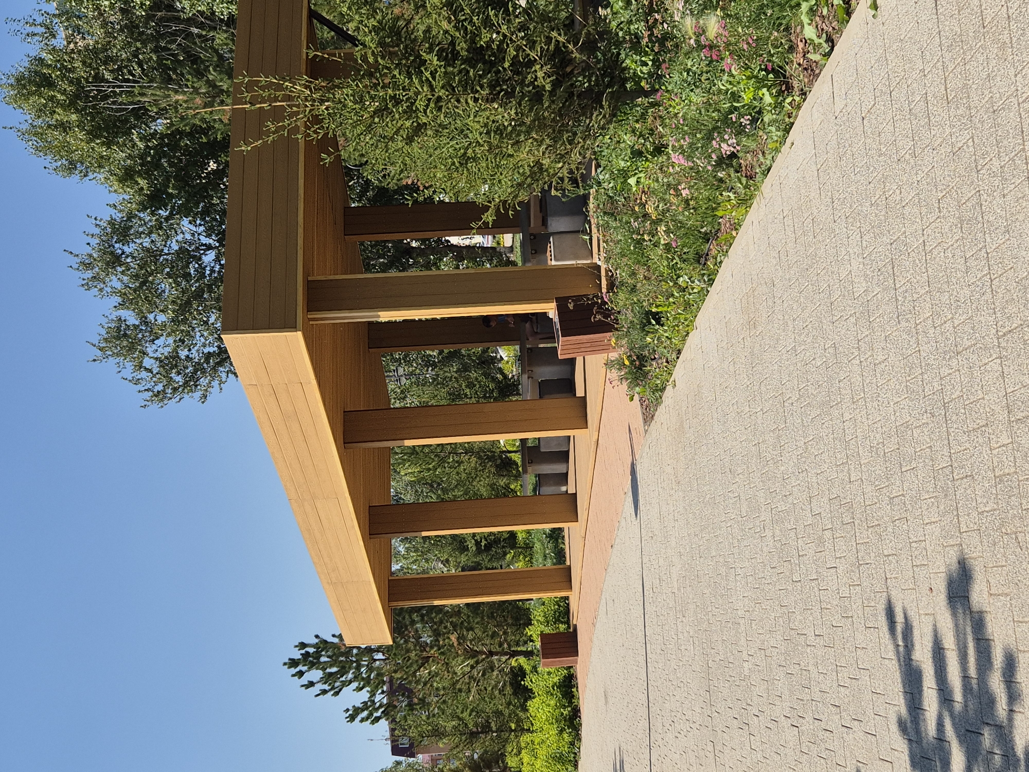 The image depicts a modern wooden gazebo situated in a park area, likely in Kokshetau, Kazakhstan, given the context. The gazebo features a flat, light brown wooden roof supported by thick, square wooden posts. Inside the gazebo, there are several concrete picnic tables and benches.
The surrounding environment is lush and green, with a variety of trees and flowering plants. A paved walkway, made of light-colored rectangular stones with a textured pattern, leads towards the gazebo, curving to the right. The walkway casts a shadow from a tree, suggesting a sunny day. A lone person, a child, is partially visible sitting on a bench inside the gazebo, facing away from the camera. To the left of the gazebo, a tall, thin wooden planter box is visible. The sky is clear and bright blue. The overall impression is one of a well-maintained public space designed for relaxation and outdoor gatherings.