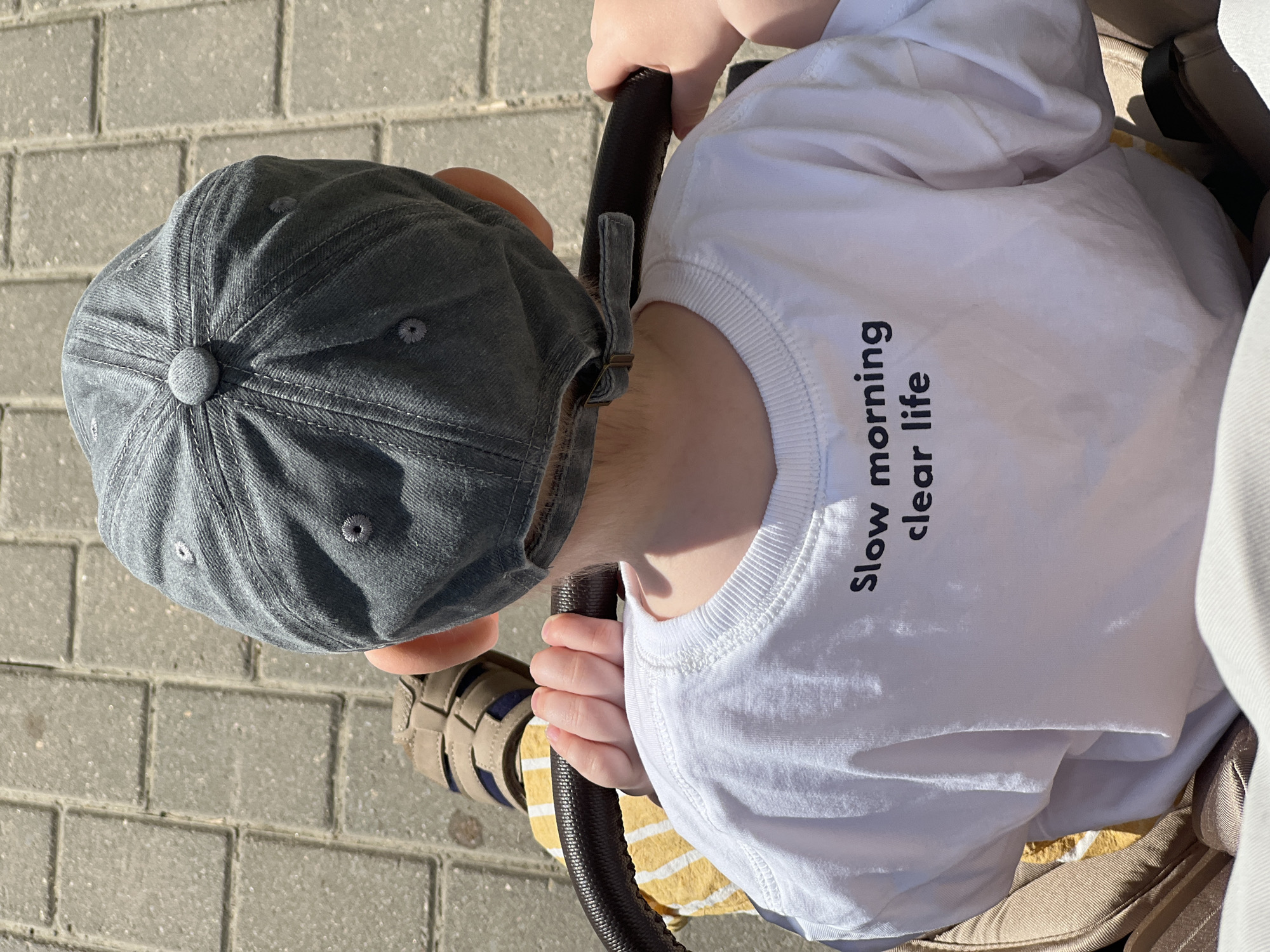 A top-down shot captures a toddler in a stroller outdoors. The child wears a light gray cap and a white shirt imprinted with "Slow morning clear life" in black text. The toddler's hands grip the stroller's handle. The stroller is tan with a black handle. One of the child's sandals is visible. The background consists of gray brick paving. The location is not explicitly identifiable as Khimki, Russia from the visual information.