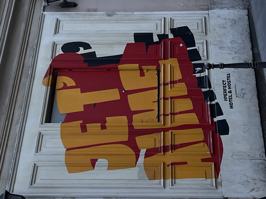 The scene displays a section of a light-colored, paneled building facade, characteristic of Haussmannian architecture, located in Paris, France, identified by embedded text as the Perfect Hotel & Hostel. A large, dynamic mural dominates the central facade. The mural features the French phrase "JE T'AIME" in bold, blocky, yellow-orange lettering with red and black layered outlines, creating a three-dimensional effect. This artwork extends across a rectangular window opening and a horizontal mail slot, integrating with the building's features. A partial black number '9' is visible in the upper right background of the mural. Below the main mural, on the lower section of the wall, smaller black text reads "#PERFECT HOTEL & HOSTEL" followed by "BY MEDELALAFOLLE". A slender, dark-colored metal bollard stands vertically in the bottom right foreground. The facade exhibits some minor surface wear and discoloration. No individuals are present in the frame.