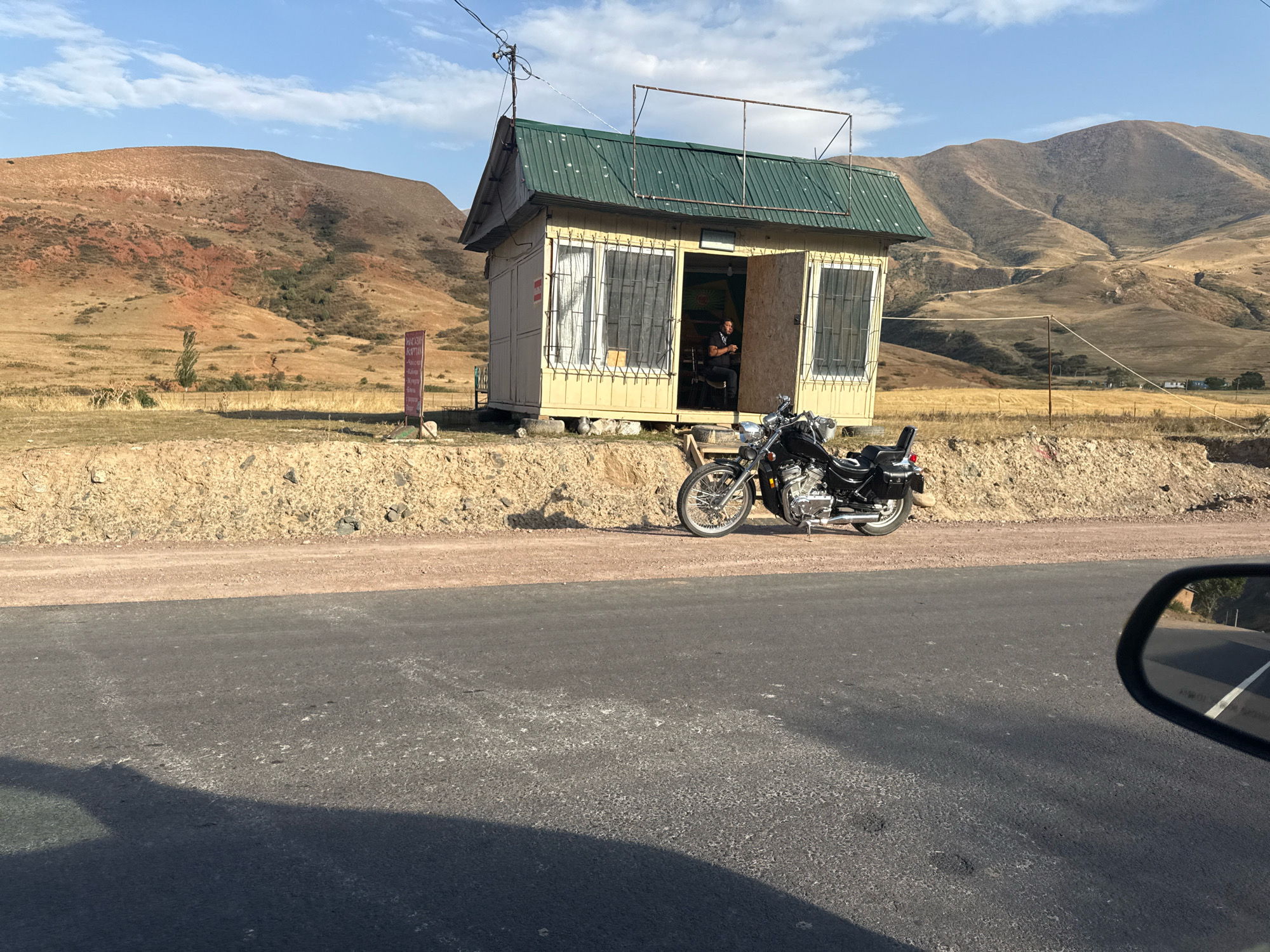 An eye-level, medium shot captures a small, light yellow, wooden structure with a green corrugated metal roof, likely a roadside kiosk or shop, situated on a dirt embankment next to a paved road. In front of the structure, a black motorcycle with chrome accents is parked on the dirt. The kiosk's entrance is open, revealing a person sitting inside on a chair. A sign with red lettering is planted in the ground to the left of the kiosk. The background features arid, hilly terrain with sparse vegetation and a clear blue sky with scattered clouds. A portion of a car's side mirror and the road from a driver's perspective are visible in the foreground. The overall setting suggests a rural or remote location, consistent with the outskirts of Arashan, Kyrgyzstan.