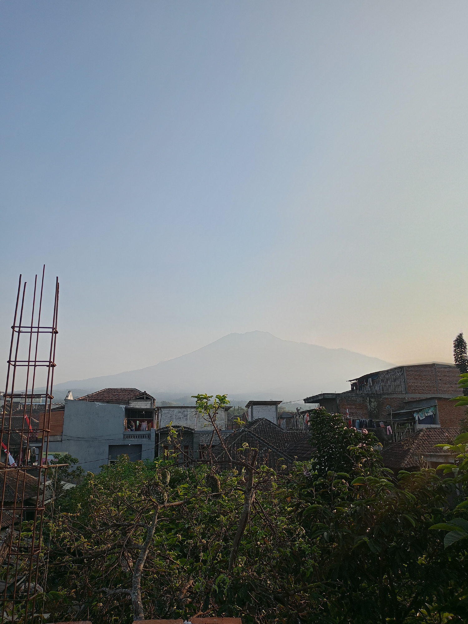 The image captures a hazy mountain range as a backdrop to a residential area in Batu, Indonesia. In the foreground, a partially constructed pillar made of rebar stands to the left. The middle ground features a cluster of houses with tiled roofs, some appearing to be multi-story. Laundry is visible hanging on clotheslines between some of the buildings. Lush green foliage, including trees and bushes, dominates the lower portion of the scene. The sky is clear and light blue, fading to a pale yellow on the horizon, suggesting either sunrise or sunset. The overall impression is of a quiet, semi-urban environment with a natural landscape.