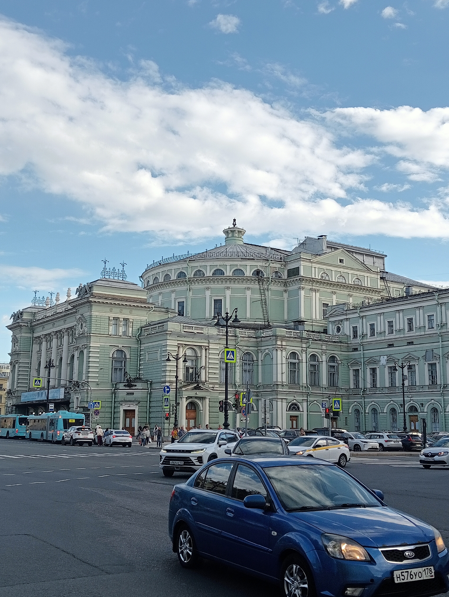 The image depicts a street scene in Saint Petersburg, Russia, under a partly cloudy sky. The focal point is a grand, light-green classical building with ornate architectural details, including columns, arched windows, and decorative friezes. The building appears to be a public institution, possibly a theater or opera house, given its scale and style.
In the foreground, several cars are driving on a paved road. A blue Kia Rio is prominently featured in the lower right corner, with its license plate visible: H576YO 178 RUS. Other vehicles include a white SUV, a white sedan, and a yellow taxi. On the left side of the image, a long, light-blue bus is stopped at what appears to be a bus stop or intersection, alongside other waiting vehicles.
People are visible on the sidewalk and crossing the street, suggesting moderate pedestrian and vehicular traffic. Pedestrian crossings and traffic signals are present, indicating an active urban environment. The weather appears to be mild, with the sun breaking through the clouds. The overall impression is that of a normal day in a European city, with a mix of historical architecture and modern transportation. The presence of a Russian license plate and the architectural style are key location cues pointing to Saint Petersburg.