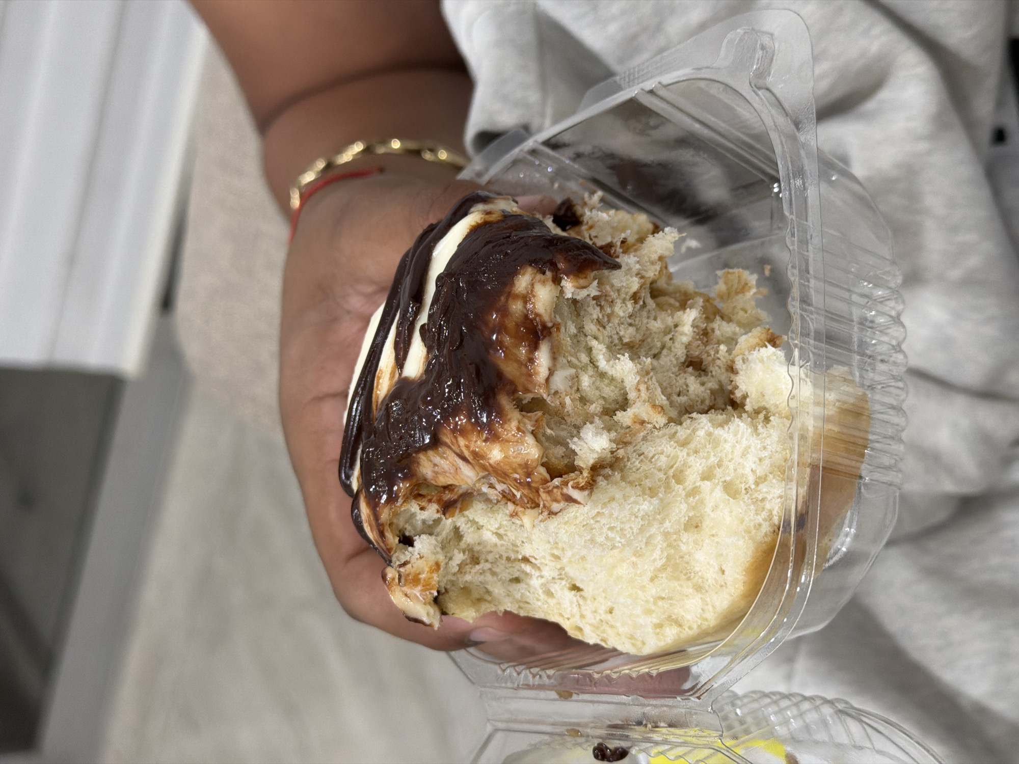 A person is holding a partially eaten, frosted pastry in a clear plastic container. The pastry is white bread with a topping of white frosting drizzled with chocolate syrup. The person's hand is holding the container and the pastry. The person is wearing a gold bracelet with a red string accent on their wrist. The person is also wearing a gray sweatshirt. The setting appears to be indoors. Without further information, the location in San Andrés Cholula, Mexico, cannot be pinpointed.