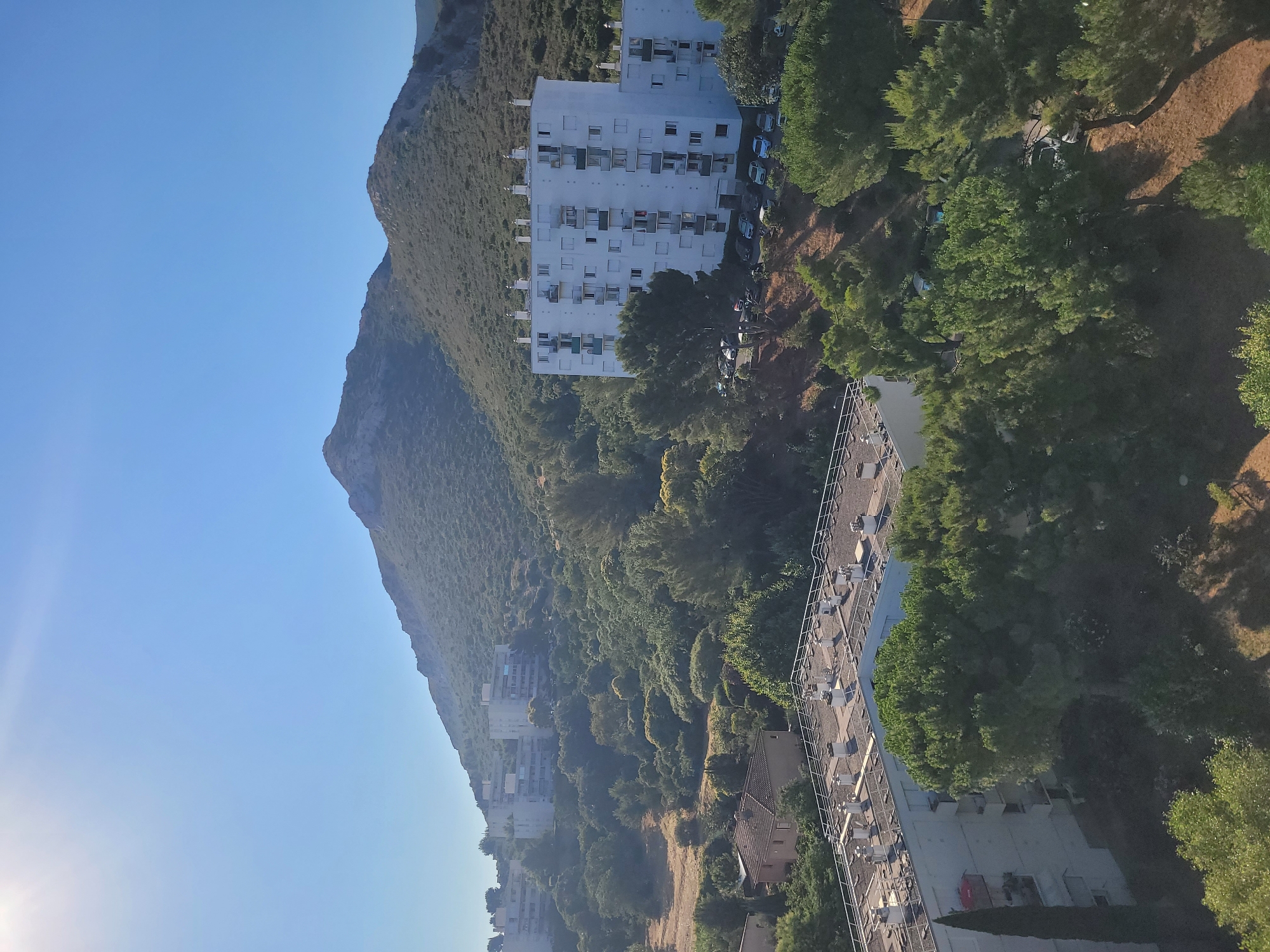 The image shows an aerial view of a residential area nestled against a green, mountainous backdrop under a clear blue sky. In the foreground, the flat roofs of several apartment buildings are visible, adorned with numerous ventilation pipes. One building, partially obscured by trees, has a narrow, tall green tree in front of it and a red object on a balcony. Further back, a large, multi-story white apartment building with many balconies and windows stands prominently, with a row of cars parked in front of it along a dirt road. The building is surrounded by lush green trees and vegetation. In the midground, more apartment buildings, appearing older and lower in profile, are visible amongst trees. A small, reddish-brown house with a tiled roof is also present. The background is dominated by a large, scrub-covered mountain that slopes gently towards the residential area. The overall scene depicts a sunny day in a semi-urban, semi-rural environment.