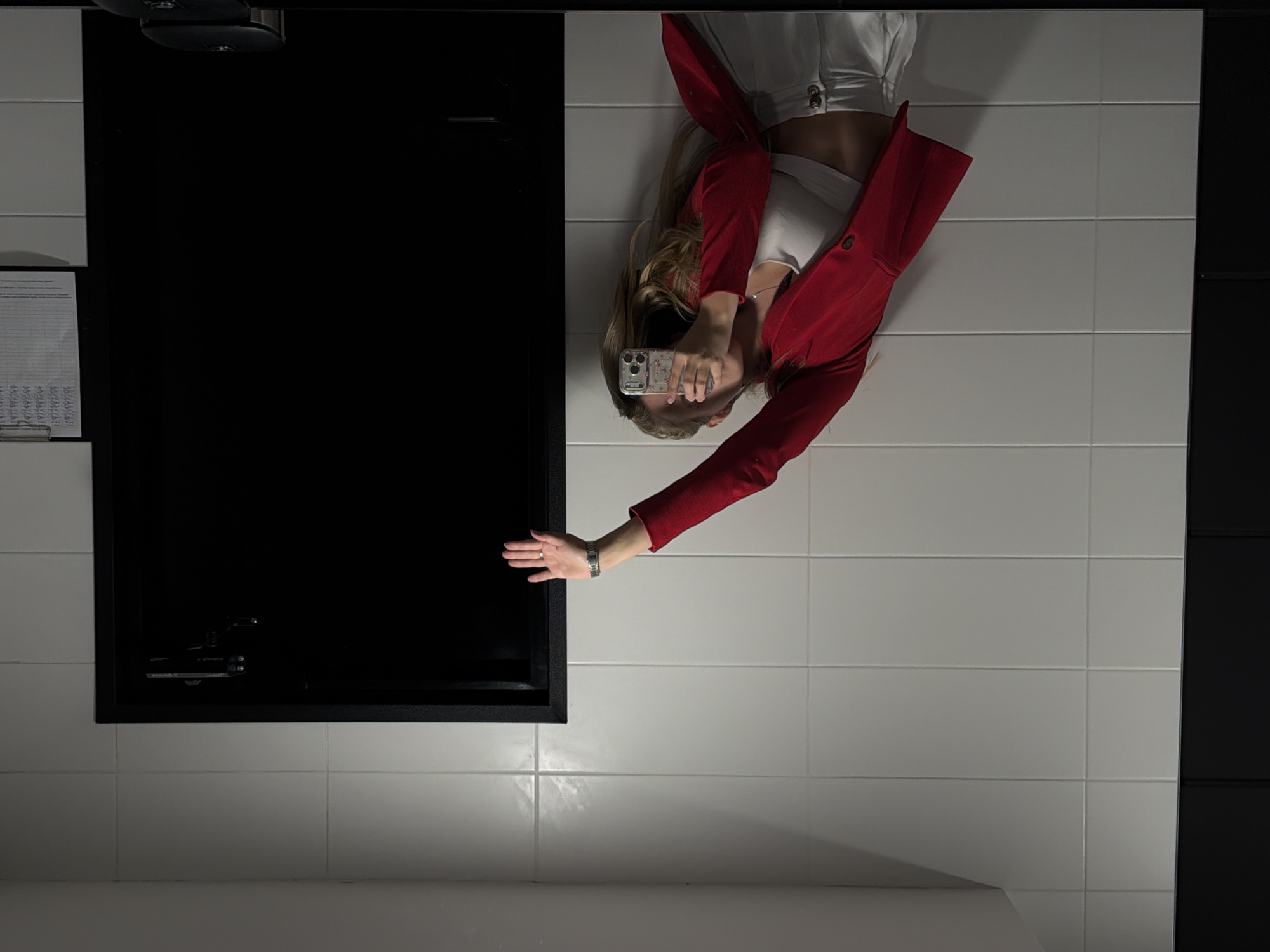A young woman, reflected in a mirror, takes a selfie in what appears to be a public restroom. She is wearing a red blazer over a white crop top and white pants. Her right arm is raised, holding a mobile phone to capture the image. The background is a tiled wall and a black door. A clipboard with papers is attached to the wall near the door. The scene is likely in Izhevsk, Russia, given the context of other images.