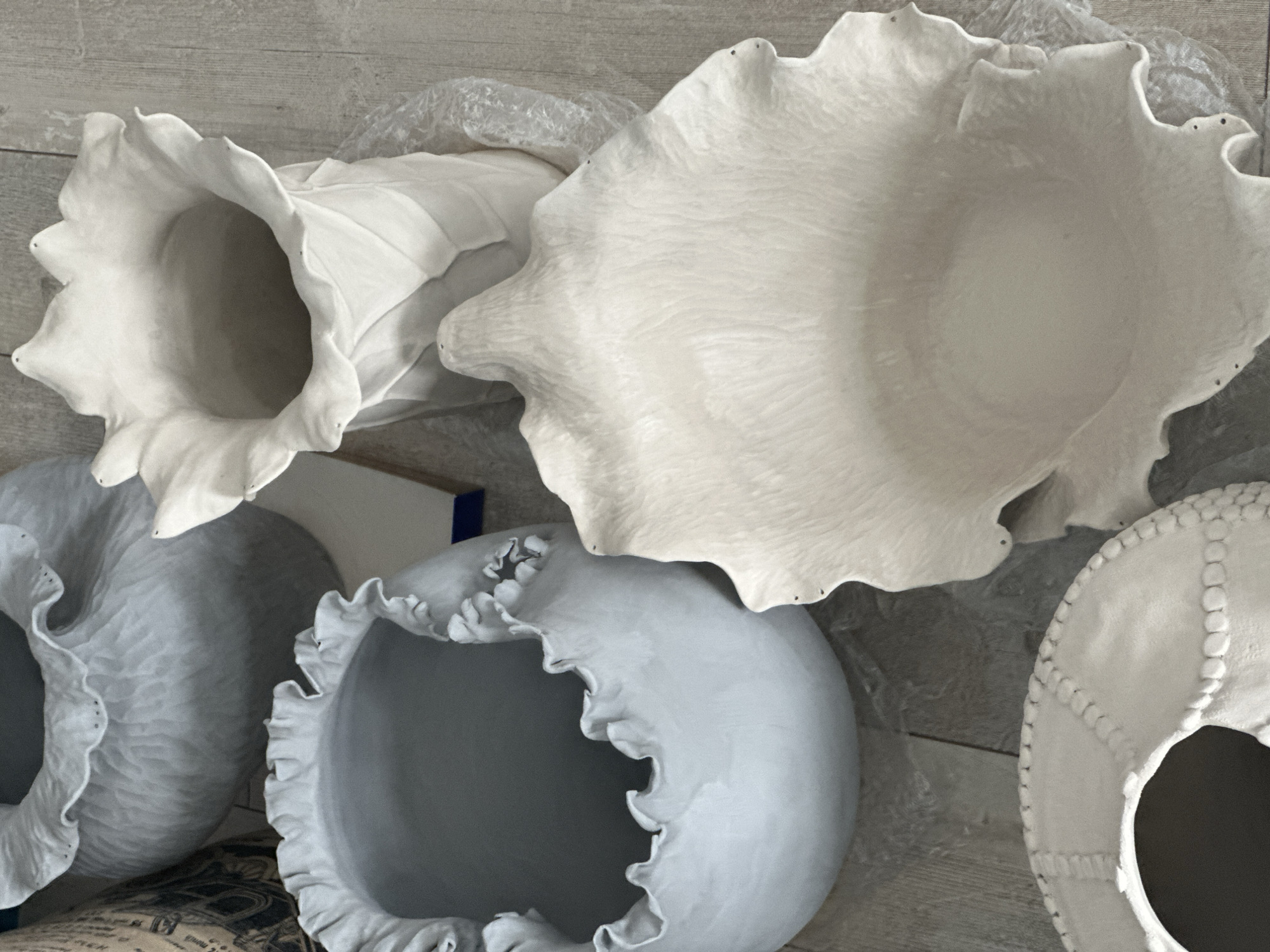 The image shows a collection of unglazed ceramic vessels, some of which appear to be vases or bowls, arranged on a wooden floor. The vessels exhibit organic, flowing shapes with ruffled edges. One large, bell-shaped vase with a flared rim is prominent in the upper right. To its left is a wider, shallower bowl with a similar ruffled edge. In the lower left, a circular, shallow dish with textured detailing around its rim is visible. The overall color palette is neutral, dominated by shades of white and gray. The scene is indoors and appears to be a studio or workspace. There are no people or animals present. The location is not specified in the image, and therefore cannot be determined to be Nevinnomyssk, Russia.