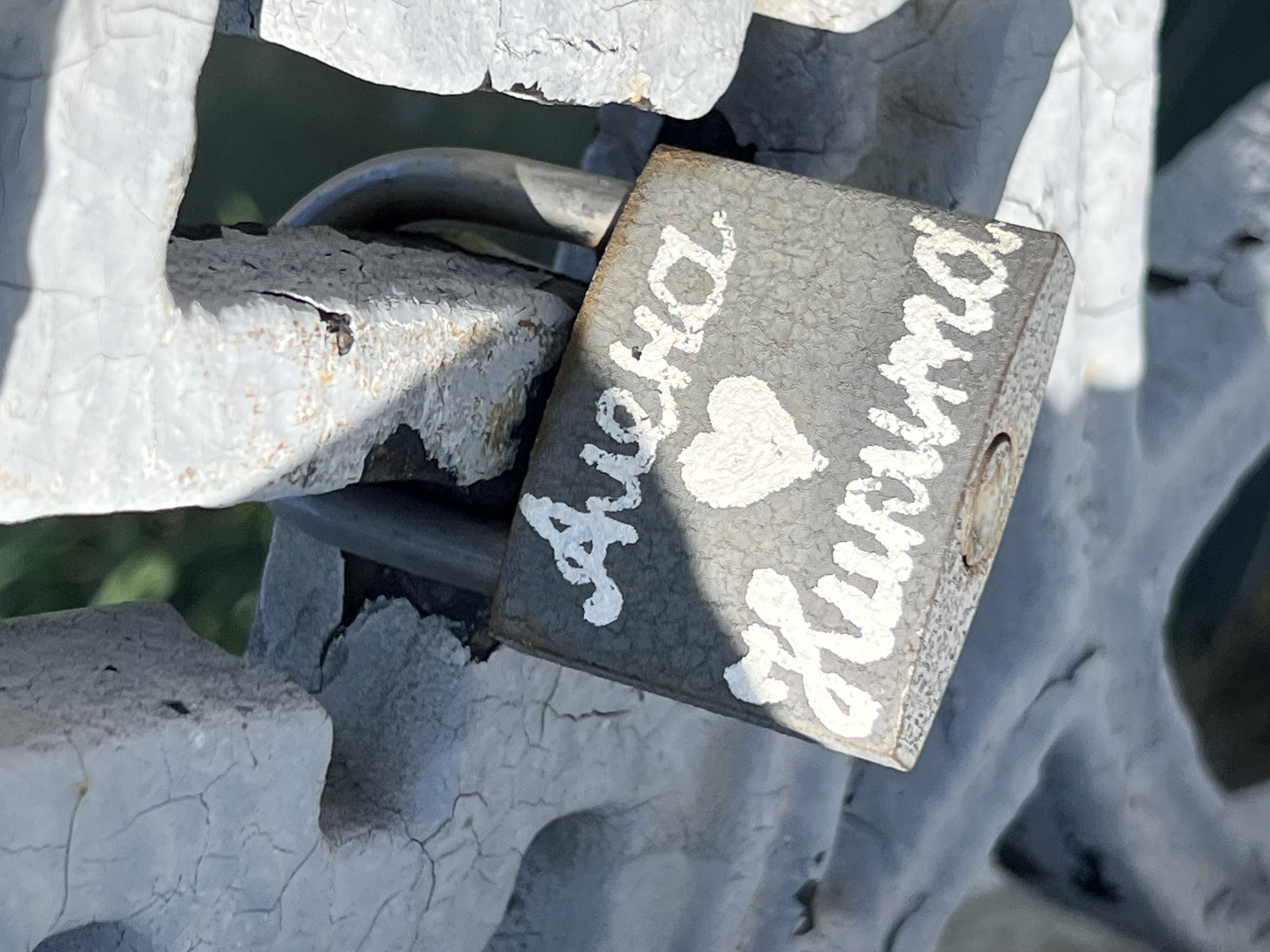 A close-up shot focuses on a gray padlock attached to a white, weathered metal structure. The padlock is adorned with white cursive lettering spelling out two names, "Aлена" and "Кристина," separated by a heart symbol. The metal structure exhibits cracked and peeling paint. The setting is Krasnodar, Russia.