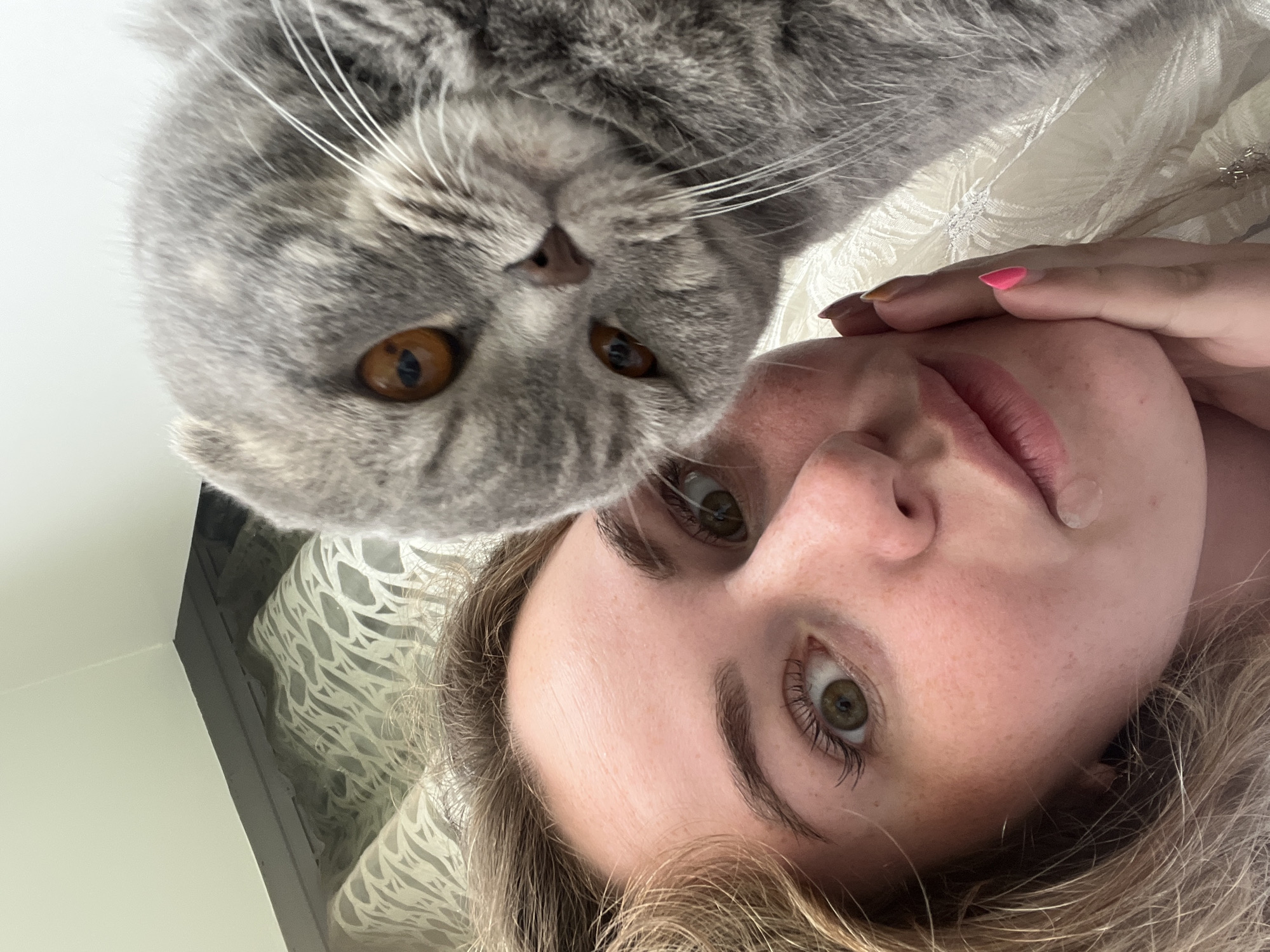 A close-up, low-angle shot captures a moment between a grey tabby Scottish Fold cat and a person. The image is framed from a perspective looking upwards, suggesting the person is lying down while the cat peers over them or is very close.
The grey Scottish Fold cat, characterized by its distinctive folded ears (partially visible) and striped fur, dominates the upper part of the frame. Its large, expressive amber-orange eyes with vertical pupils are prominent, conveying a curious or attentive gaze towards the viewer. Long, white whiskers extend from its muzzle.
The person, likely female, is seen in the lower right portion of the image. She has light-colored hair and striking green eyes. Her right hand, with light pink nail polish on one finger, rests gently near her face. A small, clear, circular patch is visible on her upper lip area. Her expression appears neutral and engaged.
The background suggests an indoor setting, likely a home, with a white patterned fabric, possibly a pillowcase or bedding, visible behind the person and cat. The lighting appears soft and natural, indicating it is daytime. The image evokes a sense of intimacy and affection between the pet and its owner. No text is visible in the image. The location context provided is Chaikovsky, Russia.