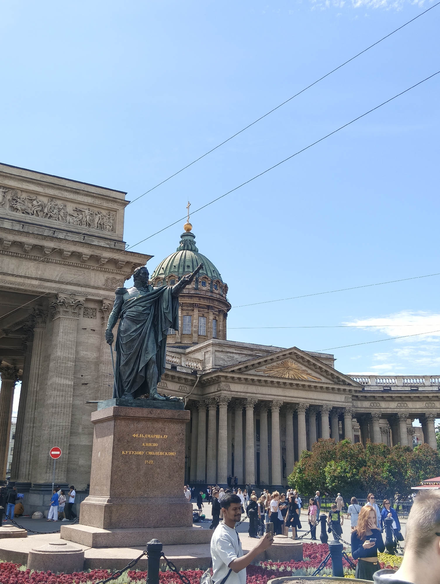 The image captures a statue of Field Marshal Kutuzov in front of Kazan Cathedral in Saint Petersburg, Russia. The statue, made of bronze, depicts Kutuzov in a military uniform, with his right arm extended forward. The base of the statue is a granite pedestal inscribed with golden Cyrillic text. Behind the statue, Kazan Cathedral is visible, characterized by its impressive colonnade and a green dome topped with a golden cross. The sky is clear blue with a few wispy clouds. In the foreground, a low chain-link fence encloses a flower bed. Several people are present in the scene, some walking and others standing. One man in the foreground is holding up a smartphone, seemingly taking a picture. The overall atmosphere is that of a public square on a sunny day.