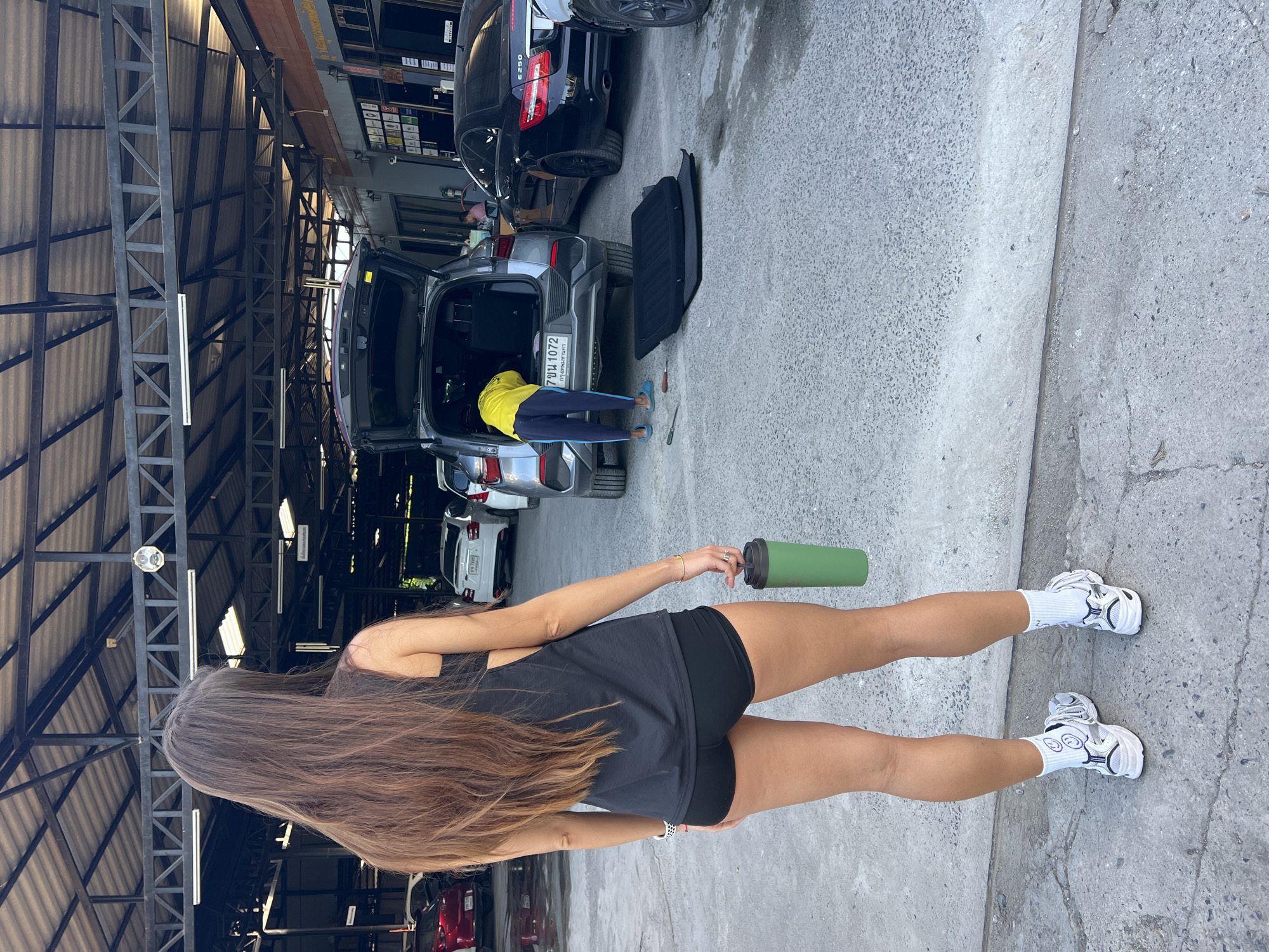 A woman with long brown hair, wearing a black tank top and black shorts, stands with her back to the camera, holding a green tumbler in her left hand. She is wearing white socks with a smiley face and white sneakers. In the background, another person wearing a yellow shirt and blue pants is working in the open trunk of a silver SUV. Next to the SUV, there is a black car and a black car mat on the ground. The vehicles are parked in what appears to be an open-air auto shop with a metal roof. The license plate on the SUV reads "7ฆณ 1072." The location is identified as Baan Lipa Noi, Thailand.