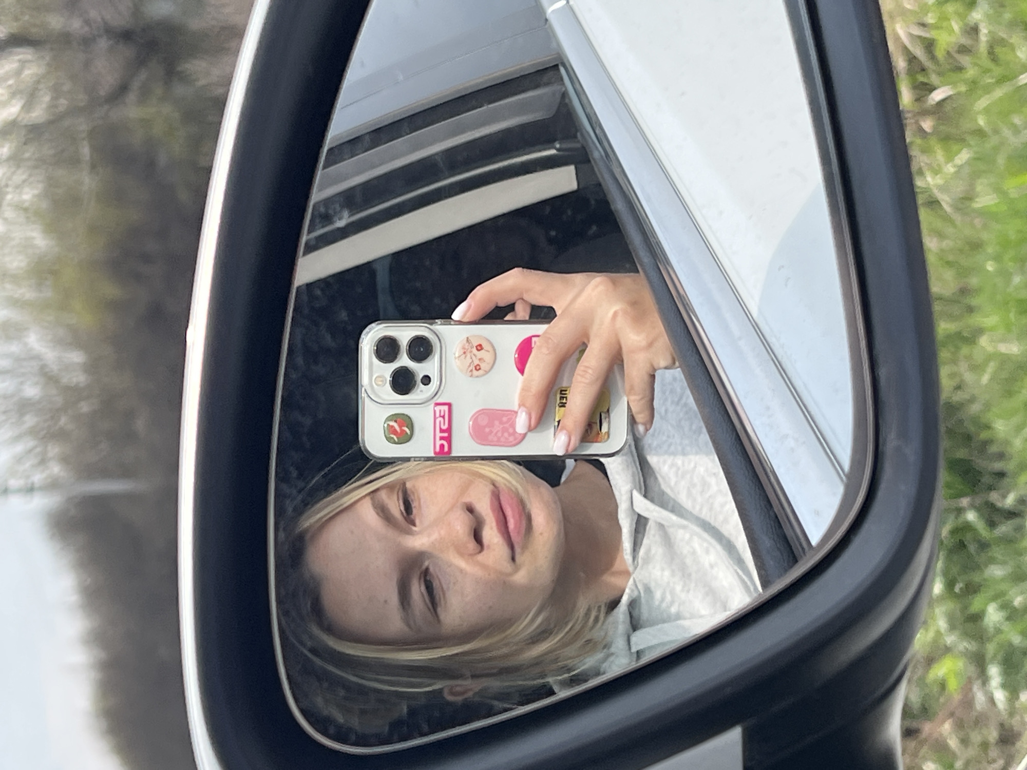 A blonde woman is captured in a selfie reflected in the side mirror of a car in Semeno-Aleksandrovka, Russia. Her face, seen from the chest up, is at the lower part of the reflection, angled slightly upwards, revealing a subtle smile and freckles. Her light blonde hair falls around her face.
Her left hand holds a smartphone, visible in the upper part of the mirror's reflection. The phone is encased in a clear cover and features several decorative elements on its back: a round popsocket with a floral pattern, a pink rectangular sticker, a yellow/gold rectangular sticker, and a red sticker displaying the white text "ERIC". Her fingernails are neatly manicured.
The mirror's reflection also shows parts of the car's interior, including a light-colored fabric of what appears to be a hoodie or sweatshirt worn by the woman, and the car's window frame. Outside the car, visible in the mirror and faintly in the background, is a blurred natural environment with green foliage and trees, suggesting a sunny daytime setting in an outdoor area.