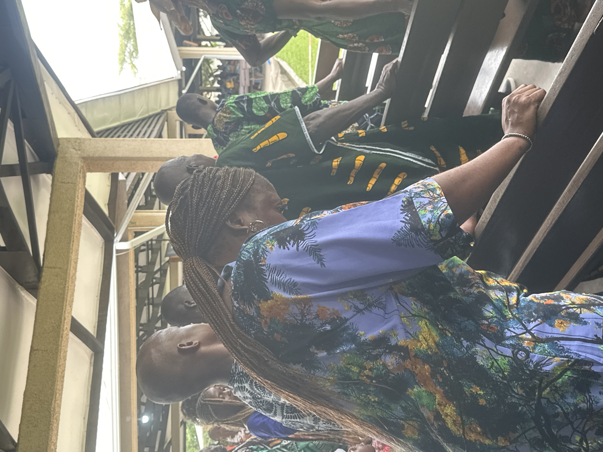 In Itoga, Nigeria, a group of individuals are gathered outdoors, possibly at a community event or religious service. The scene is characterized by a covered outdoor seating area with wooden benches. The focus is on several people standing and interacting, their attire predominantly featuring vibrant, patterned fabrics, possibly traditional African clothing. One woman in the foreground wears a distinctive purple garment with a detailed forest or tree motif. Her braided hair extends down her back. Other individuals are visible in the background, some seated on the benches, while others stand. The overall environment suggests a casual yet possibly formal gathering, with hints of natural light filtering through the covered structure. The setting appears to be open and airy.