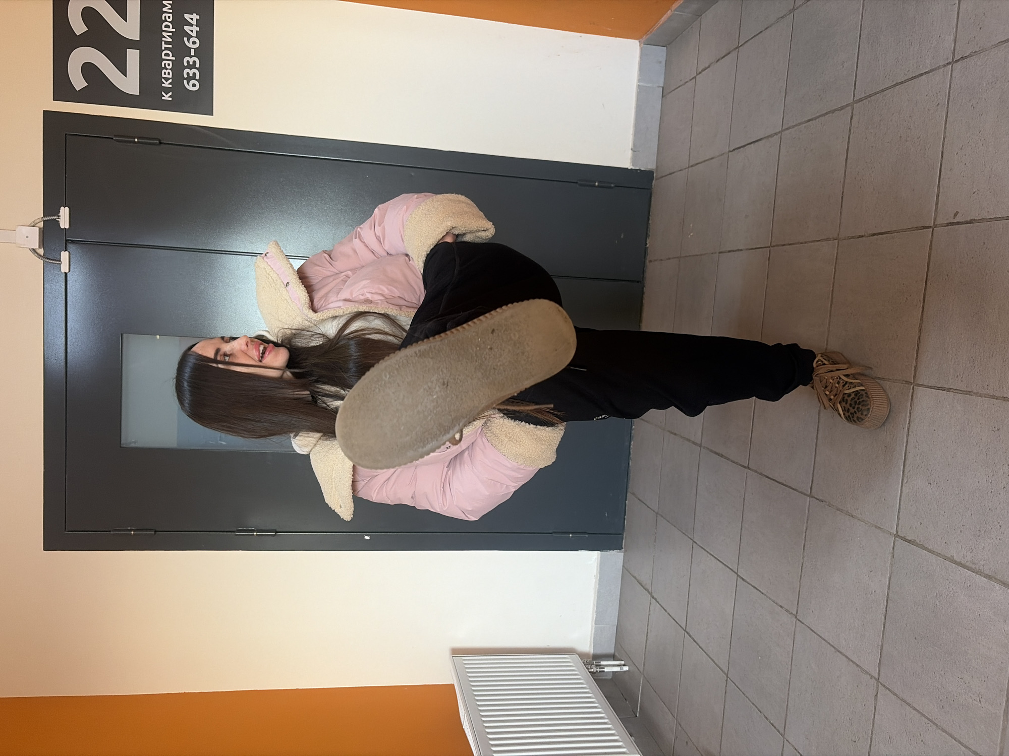 A young woman stands in a hallway in Saint Petersburg, Russia, extending her left leg forward, showing the sole of her shoe. She has long dark hair and wears a pink jacket with a furry collar, black pants, and tan sneakers. Behind her is a dark grey door with a sign reading "22 к квартирам 633-644". The walls are painted white with an orange stripe at the bottom, and the floor is tiled. A white radiator is visible in the lower part of the frame.
