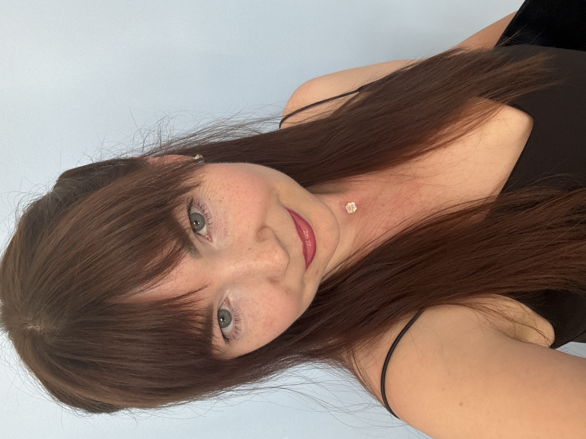 A close-up portrait features an adult individual, appearing female, centered against a plain light blue wall. The individual has long, straight, dark brown hair with a fringe covering the forehead, light-colored eyes (appearing blue-green or grey-green), and a fair complexion adorned with numerous freckles across the nose and cheeks. They wear a black spaghetti strap top. A small, iridescent, cube-shaped pendant on a thin chain is visible at the base of their throat, and a small, clear stud earring is in their right ear. A subtle smile is present, and the lips are colored with a berry-toned glossy lipstick. The individual looks directly towards the camera. The lighting is soft and even, suggesting an indoor setting. This scene is located in the city of Chaikovsky, Russia.