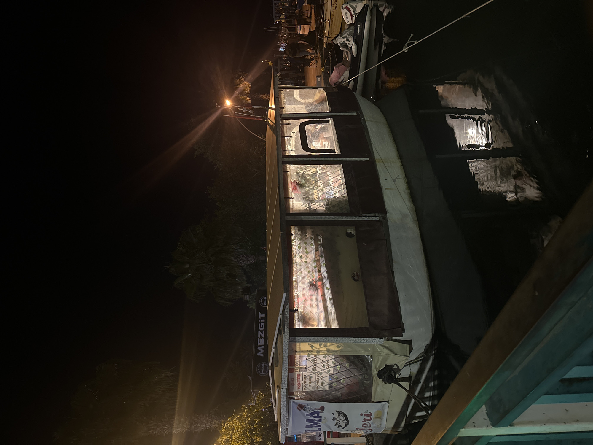 The image shows a boat restaurant named MEZGIT docked at night in Fethiye, Turkey. The boat is illuminated from the inside, revealing tables and chairs. Menus and advertisements are displayed near the entrance. The water reflects the boat's lights, and a street lamp shines in the background. A crowd of people is visible farther down the pier where the boat is docked.