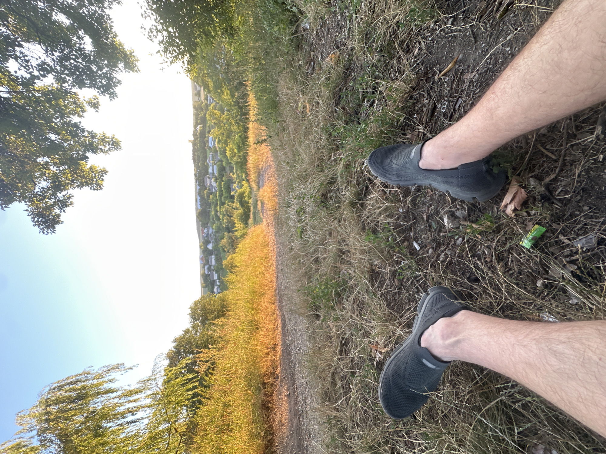 In Makiv, Ukraine, a close-up, low-angle shot captures a person's legs and feet, clad in dark, slip-on athletic shoes, resting on dry, golden grass. The scene is set outdoors, with a path made of gravel and dirt leading away from the viewer. In the background, a gentle slope descends into a valley dotted with houses and trees, suggesting a rural or semi-rural setting on the outskirts of a settlement. The setting sun casts a warm, golden light on the dry vegetation, creating a serene, late-afternoon atmosphere. The sky is a clear, bright blue, with some tree branches framing the upper portion of the image. A small, green candy wrapper is visible on the ground near the person's feet.