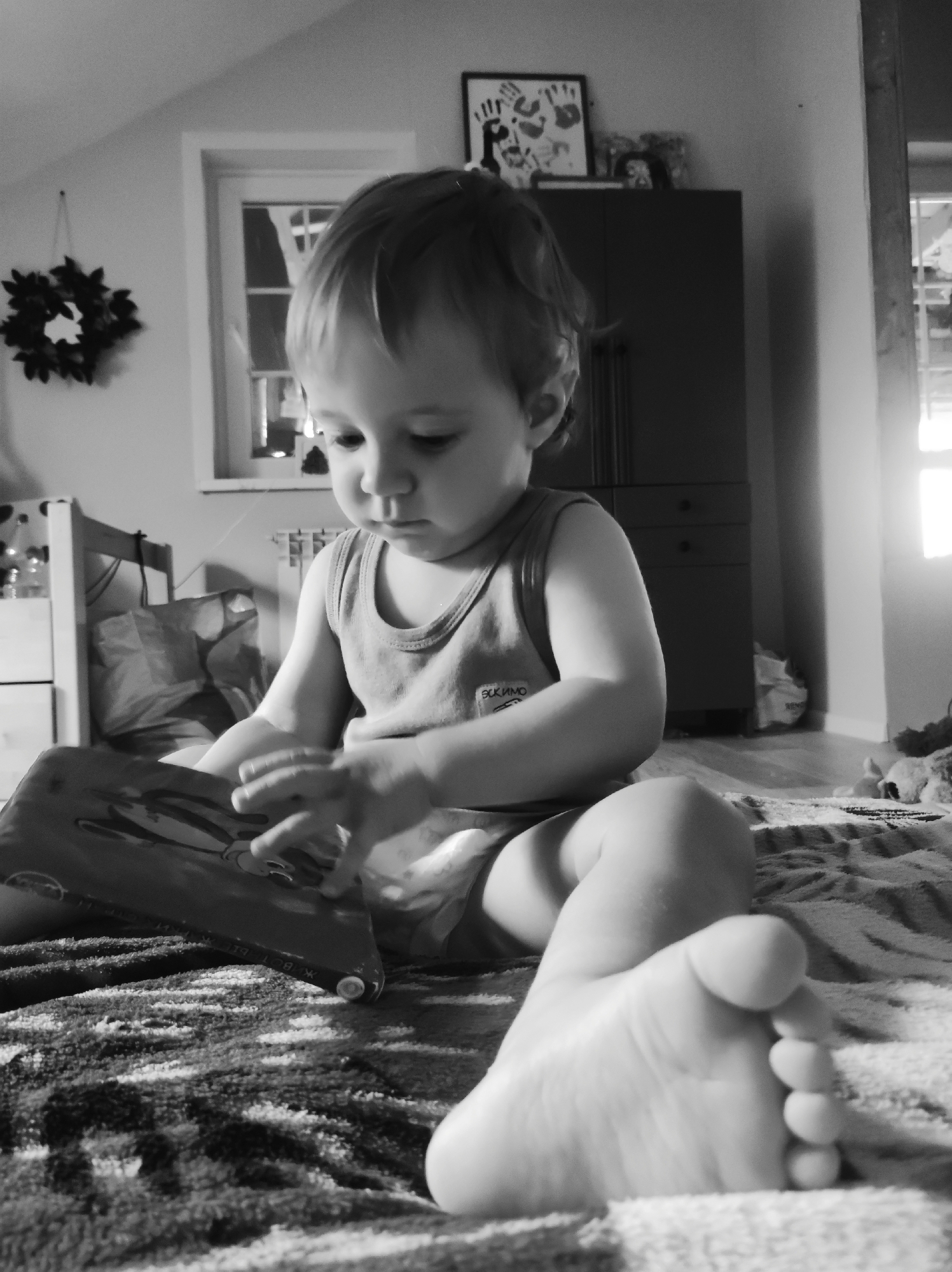 This black and white image depicts a toddler, gender indeterminate, seated on a textured blanket or rug in an interior room located in the city of Unknown, Unknown. The child is positioned centrally, looking downwards, and engaged with an open book held by both hands. They are dressed in a light-colored sleeveless undershirt, which features the Cyrillic text "Эскимо" on the left chest area, and light-colored shorts or a diaper. Both feet are bare, with the right foot prominently visible in the lower foreground.
The background on the left features a white-framed multi-pane window, allowing natural light into the room; a dark, circular decorative wreath-like item hangs on the wall to its left. A radiator is partially visible below the window, and the wall above the window indicates a sloped ceiling. The background on the right presents a dark wooden cabinet or wardrobe with multiple doors and drawers. A framed piece of artwork, consistent with child's handprints, and other small objects are displayed on top of this furniture. To the far right, a vertical wooden structural beam or doorframe is present, along with an additional source of natural light. The floor under the blanket appears to be wood.