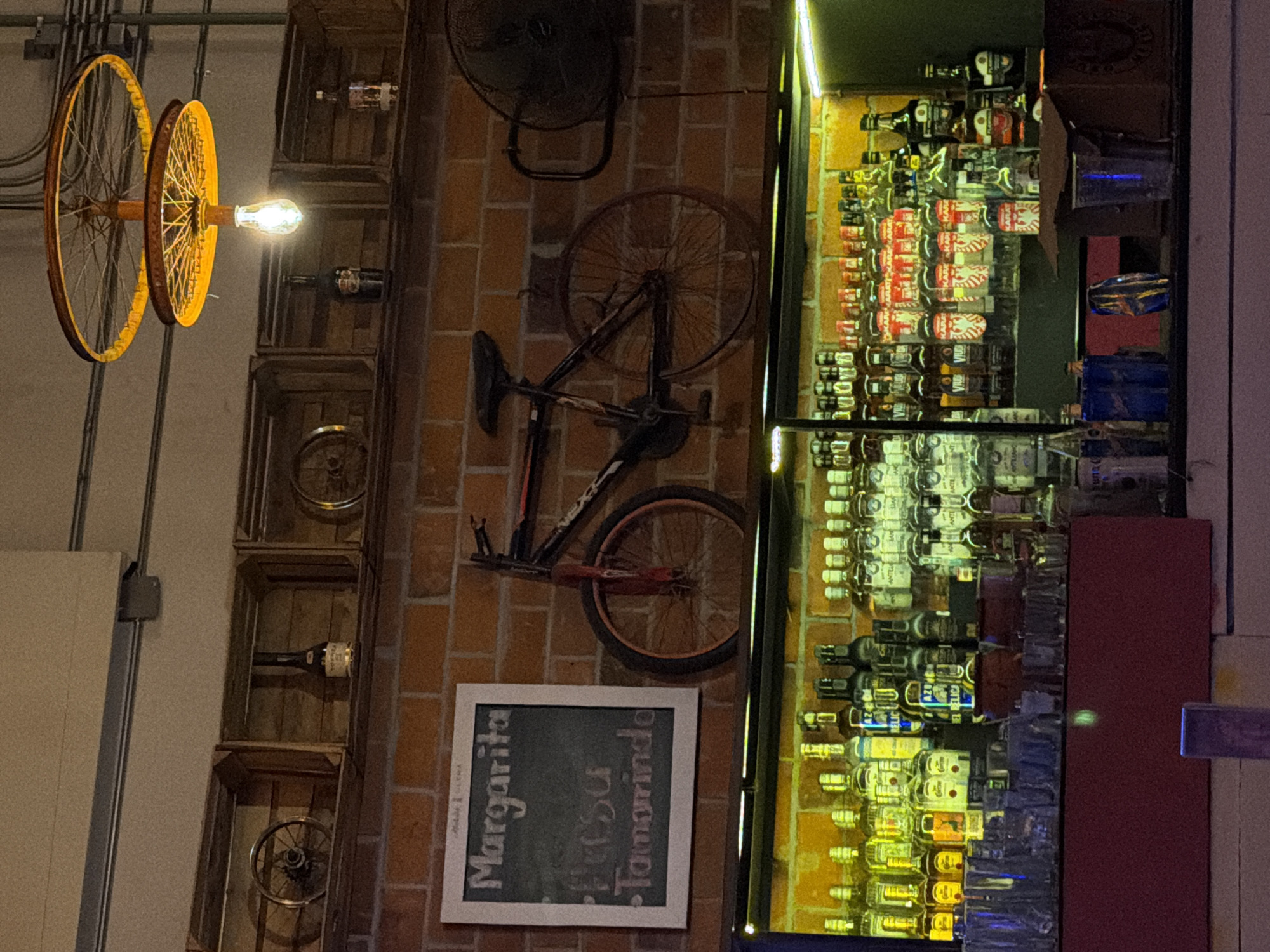 The image displays the interior of a bar or restaurant located in Puebla, Mexico. The scene is static, with no individuals present.
In the foreground, a maroon-colored bar counter is equipped with numerous clear drinking glasses, a few blue and yellow containers, and a brown cardboard box on its surface. Behind this counter, an illuminated shelving unit showcases rows of various liquor bottles, predominantly clear glass with assorted labels, including partially visible brands like "Karat." These bottles are arranged against an exposed brick wall, bathed in the greenish-yellow light from an unseen source beneath the shelves.
Above the liquor display, the brick wall continues as a prominent feature. Mounted horizontally on this wall is a dark-framed bicycle with a distinct red front fork and wheel. To the left of the bicycle, a white-framed chalkboard displays handwritten text in white chalk, listing "Margarita," "Fresa" (strawberry), and "Tamarindo" as options. To the right of the bicycle, a black industrial-style fan is affixed to the wall.
Higher on the wall, a series of wooden crates are utilized as shelves, containing decorative items such as additional bicycle wheel components and various bottles, including one dark bottle resembling a cream liqueur. Suspended from the light-colored ceiling, which features visible grey conduit pipes, is a unique chandelier crafted from two inverted bicycle wheels, positioned one above the other. An exposed incandescent light bulb hangs centrally from the lower wheel, emitting a warm, downward-focused glow.