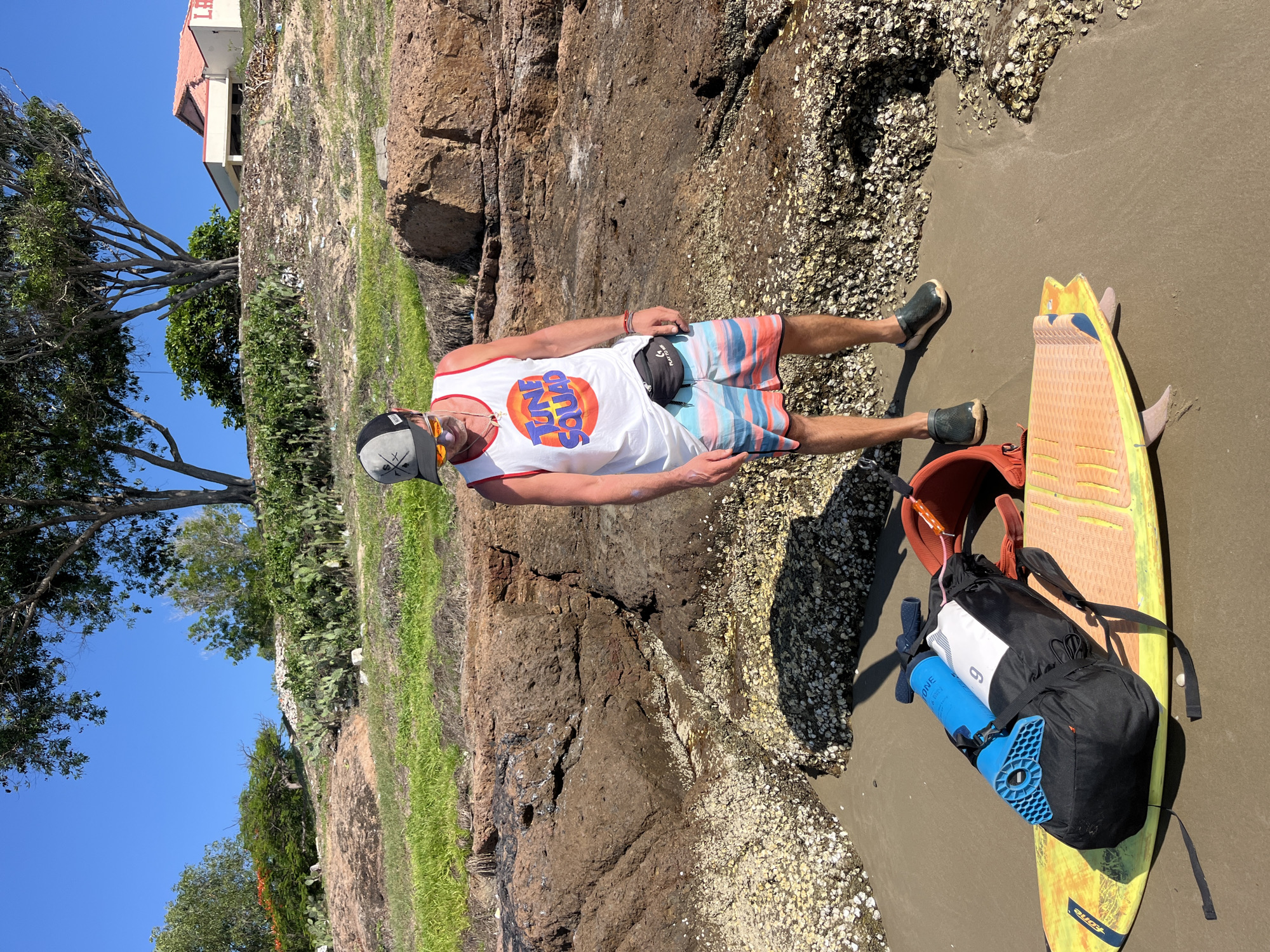 In the city of Mũi Né, Vietnam, a man stands on a sandy beach near rocky outcrops. He is wearing a white "Tune Squad" tank top, striped shorts, a baseball cap, sunglasses, and dark clogs. A black fanny pack is worn around his waist. In the foreground, a yellow surfboard lies on the sand, with a black backpack, an orange bag, and a blue object placed on top of it. The background shows a grassy hillside with trees, cacti, and a building with a red roof. The sky is clear and blue.