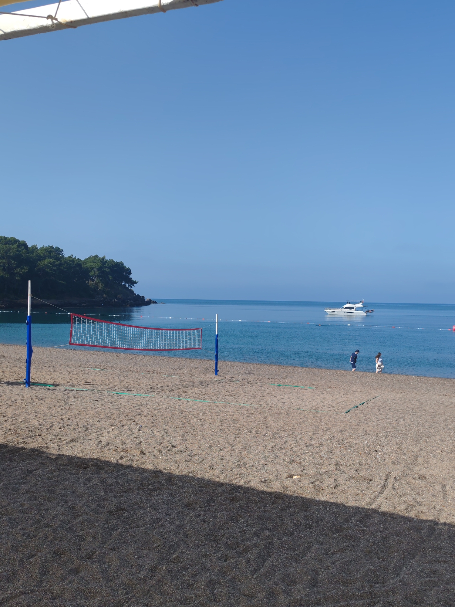 The image shows a beach scene in Alanya, Turkey. A volleyball net stands on the sandy beach, with a coastline to the left topped with trees. The sea is calm and blue with a yacht in the distance. Two people stand near the shoreline. A shadow from an unknown object is cast onto the foreground sand. The sky is clear and blue.
