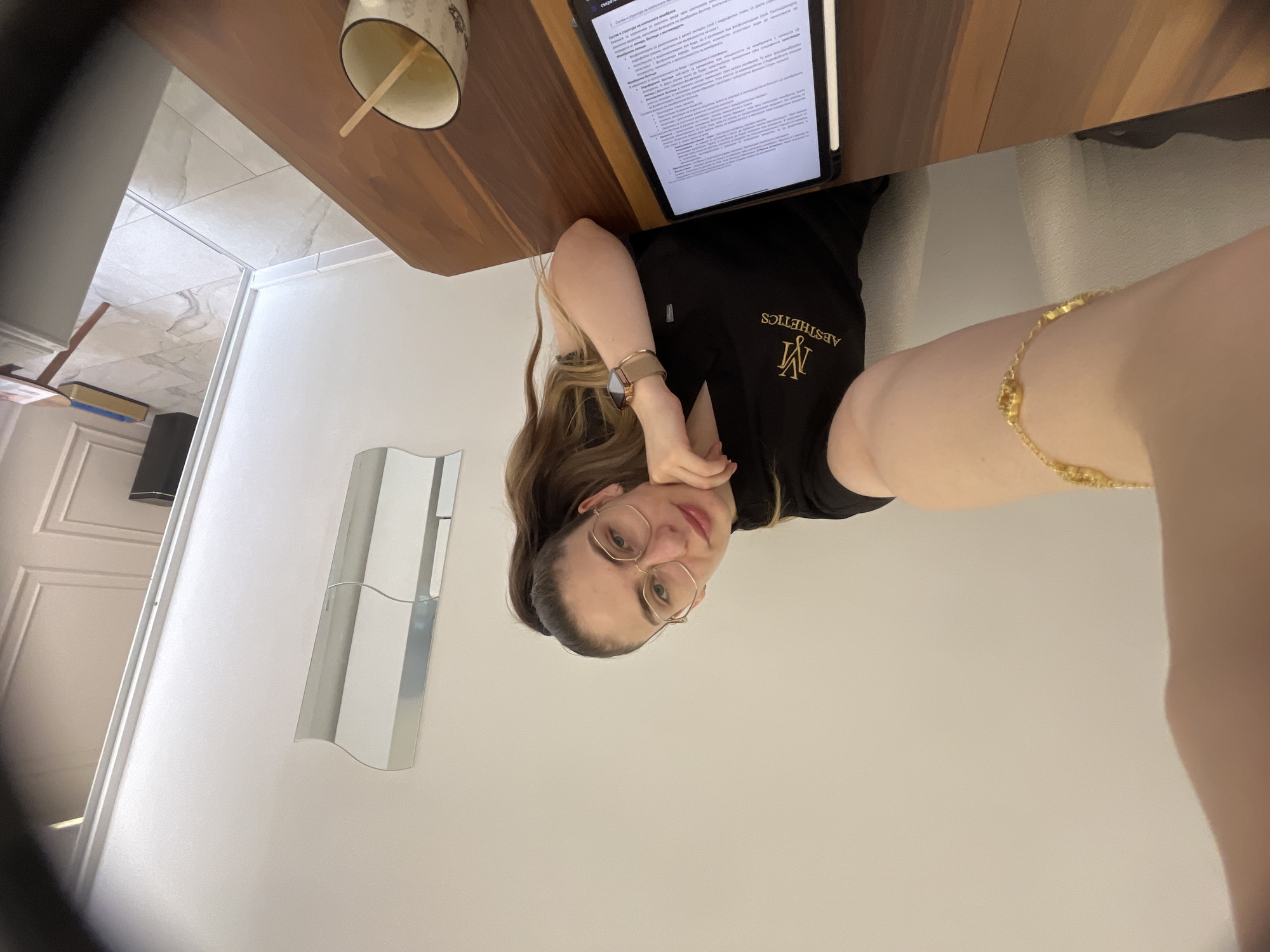 A woman with long brown hair and glasses sits at a wooden table, looking at the camera. She is wearing a black scrub top with an "M AESTHETICS" logo. Her left hand is resting on her chin, and her right arm is extended towards the camera, holding her phone. On the table in front of her is a tablet displaying a document and a mug with a wooden stir stick. The room is sparsely decorated with white walls and a modern mirror on the wall. The floor is tiled.