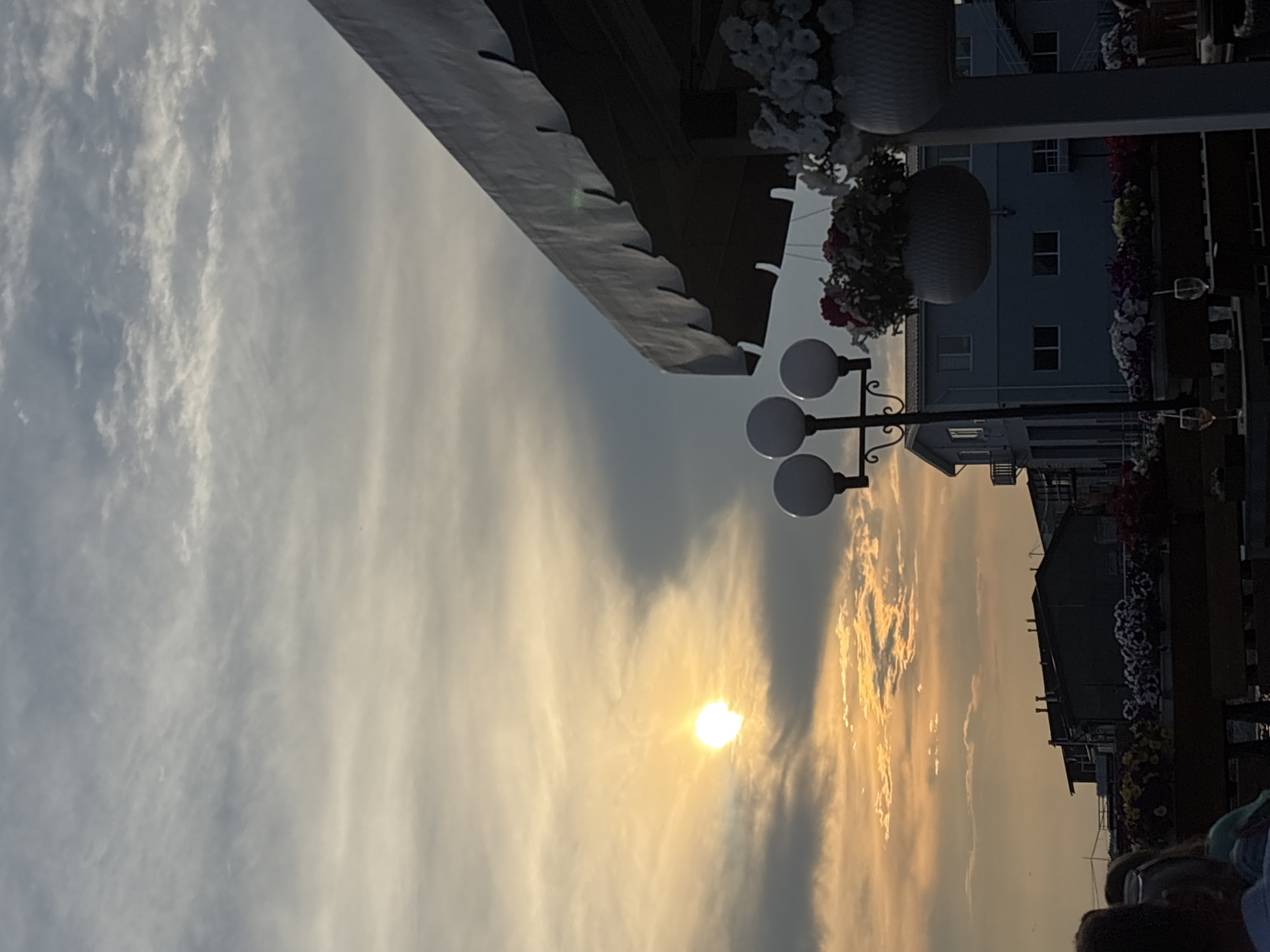 The image depicts a sunset over Tolyatti, Russia. The sun is a bright orb partially obscured by clouds, casting a warm glow across the sky. In the foreground, a portion of an awning from a cafe or restaurant is visible, along with hanging flower baskets and outdoor lighting fixtures. In the mid-ground, several buildings with dark rooftops can be seen, suggesting a developed area. The overall atmosphere is serene and evocative of an evening in a coastal or lakeside city.
