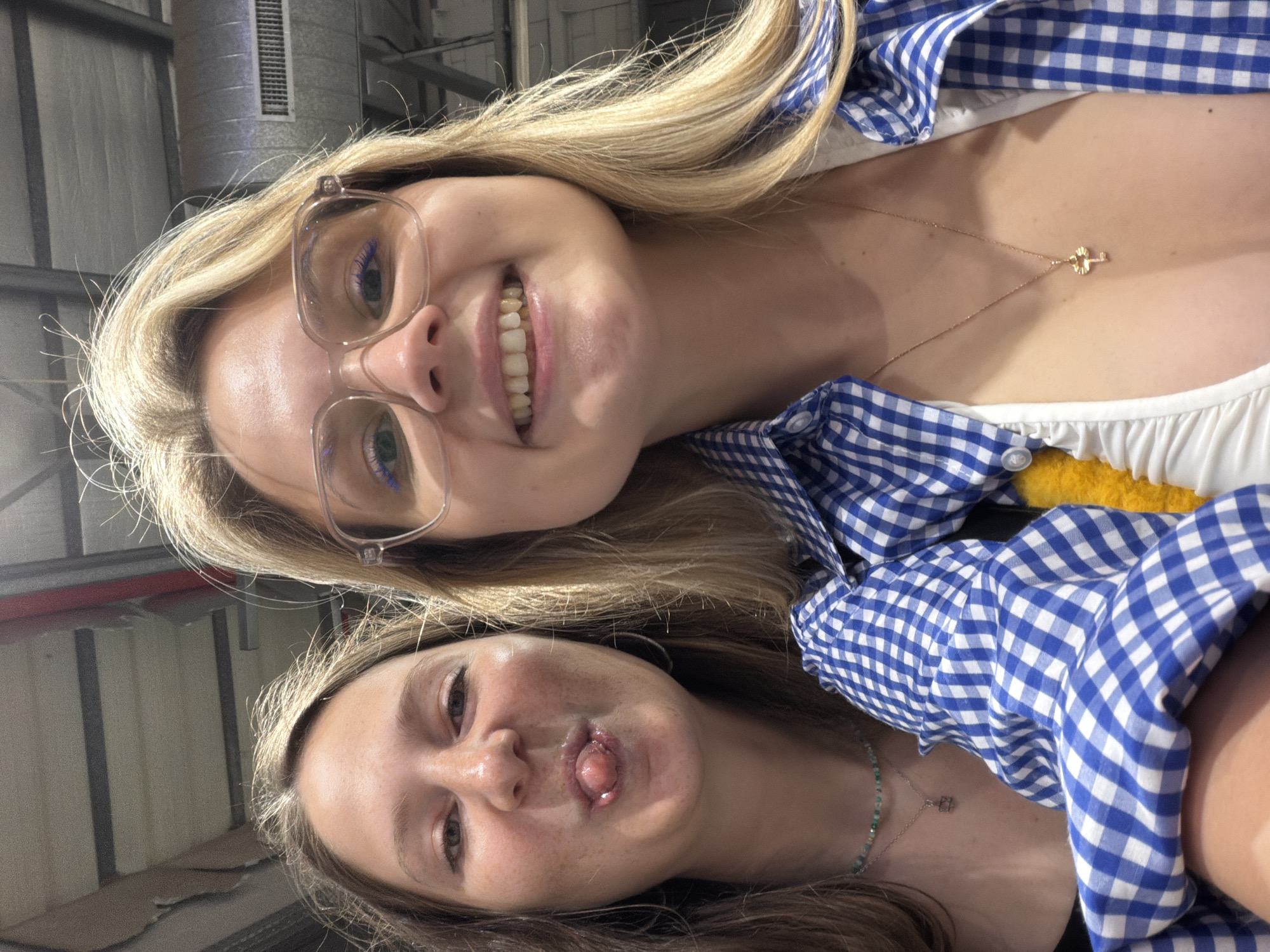 Two women are posing for a selfie. The woman on top has long blonde hair, glasses, and a blue and white checkered shirt. She is smiling and wearing a necklace with a key pendant. The woman on the bottom has long brown hair and is sticking her tongue out. She is also wearing a necklace. The background appears to be an industrial setting, possibly indoors. The specific location within Bat Yam, Israel, is not discernible from the image.