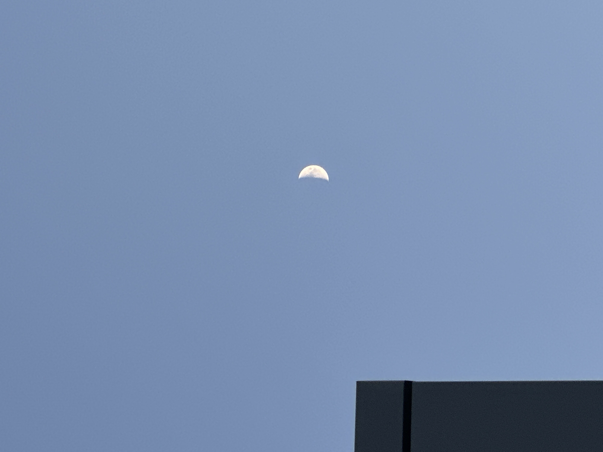 The image shows a half moon in a blue sky above a dark, flat rooftop. The composition is simple, with the moon centered in the upper part of the frame and the rooftop occupying the bottom right corner. There are no people visible. The image does not provide enough information to confirm its location in Chaikovsky, Russia.