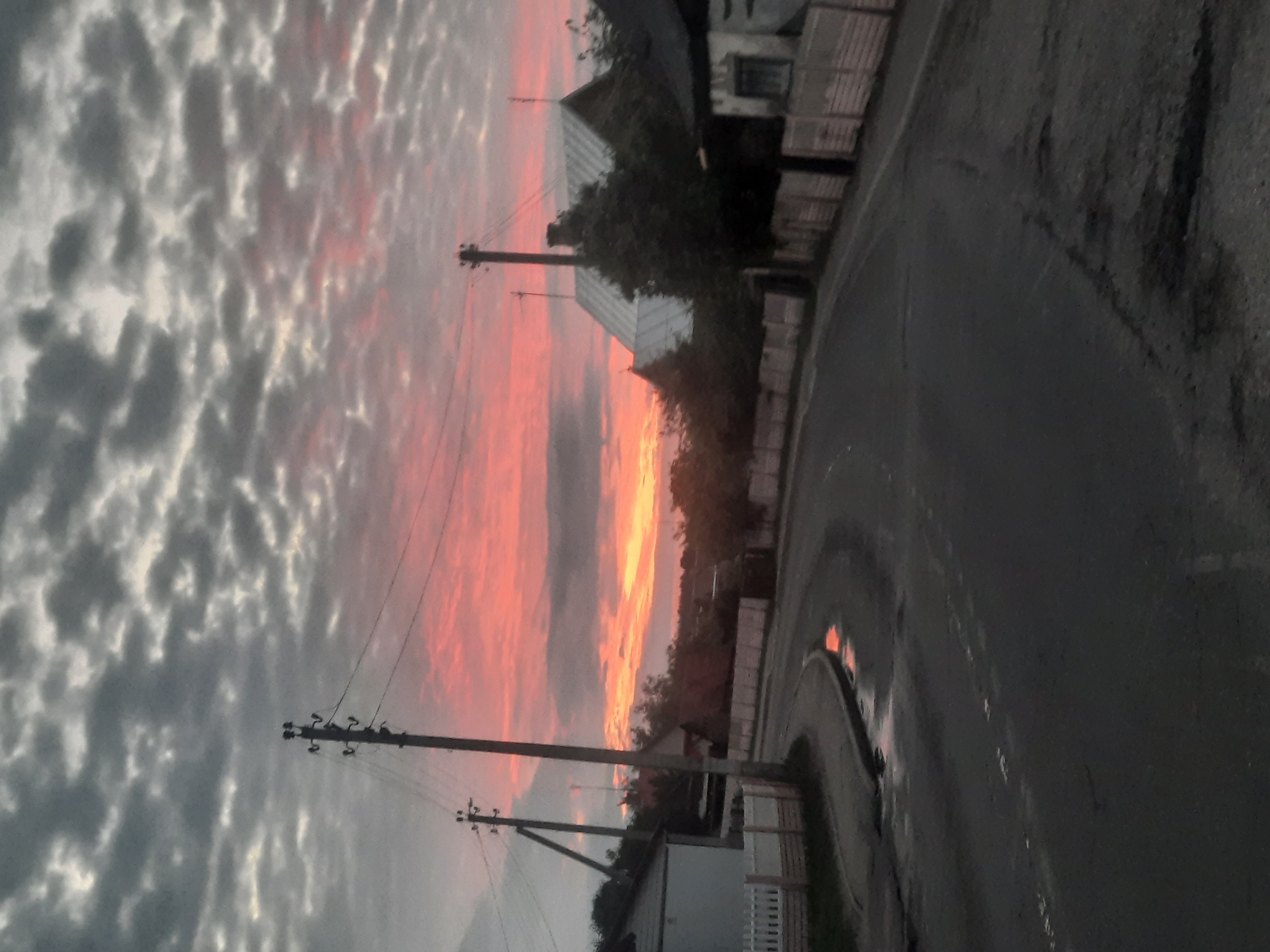 The image captures a rural scene in the city of Parychcha, Belarus, at what appears to be dusk or dawn. A paved road curves through a village, bordered by wooden fences and houses. The sky is filled with dramatic clouds, colored with hues of pink, orange, and grey from the setting or rising sun. Electrical poles with wires run along the side of the road. A puddle on the road reflects the colorful sky. The atmosphere is serene, with no people or animals visible. The overall impression is one of quiet rural life under a striking sky.