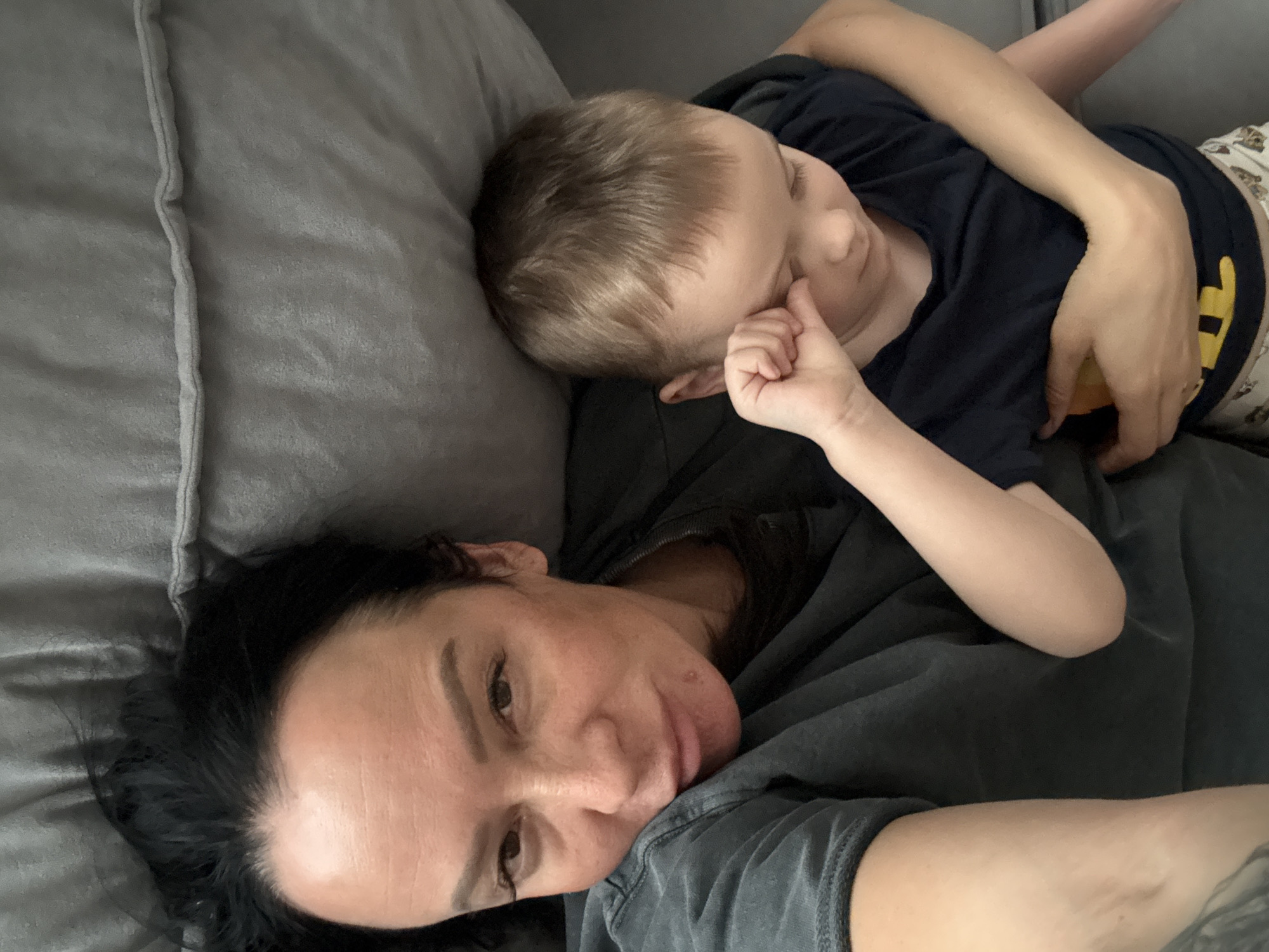 An overhead view captures an adult woman and a young child resting together on a grey, upholstered surface, likely a sofa or bed. The scene is set indoors in Chaikovsky, Russia.
The woman is in the foreground, lying on her back or side, with her head turned towards the camera. She has dark hair, visible around her head, and is wearing a dark, possibly grey, t-shirt. Her expression is calm, and she appears to be looking directly at the camera.
Behind and above the woman, a young child is nestled against her. The child, with light brown hair, is lying on their side, facing away from the camera and towards the woman's shoulder area. They are wearing a dark blue t-shirt and light-colored, patterned shorts. One of the child's hands is near their mouth, suggesting comfort or sleep. The child's posture indicates a deep state of relaxation or sleep, snuggled close to the woman.
The overall atmosphere of the image is one of peace, intimacy, and comfort, highlighting a tender moment of rest or affection between the two individuals. The fabric of the couch or bed has a soft, textured appearance. There is no visible text in the image.