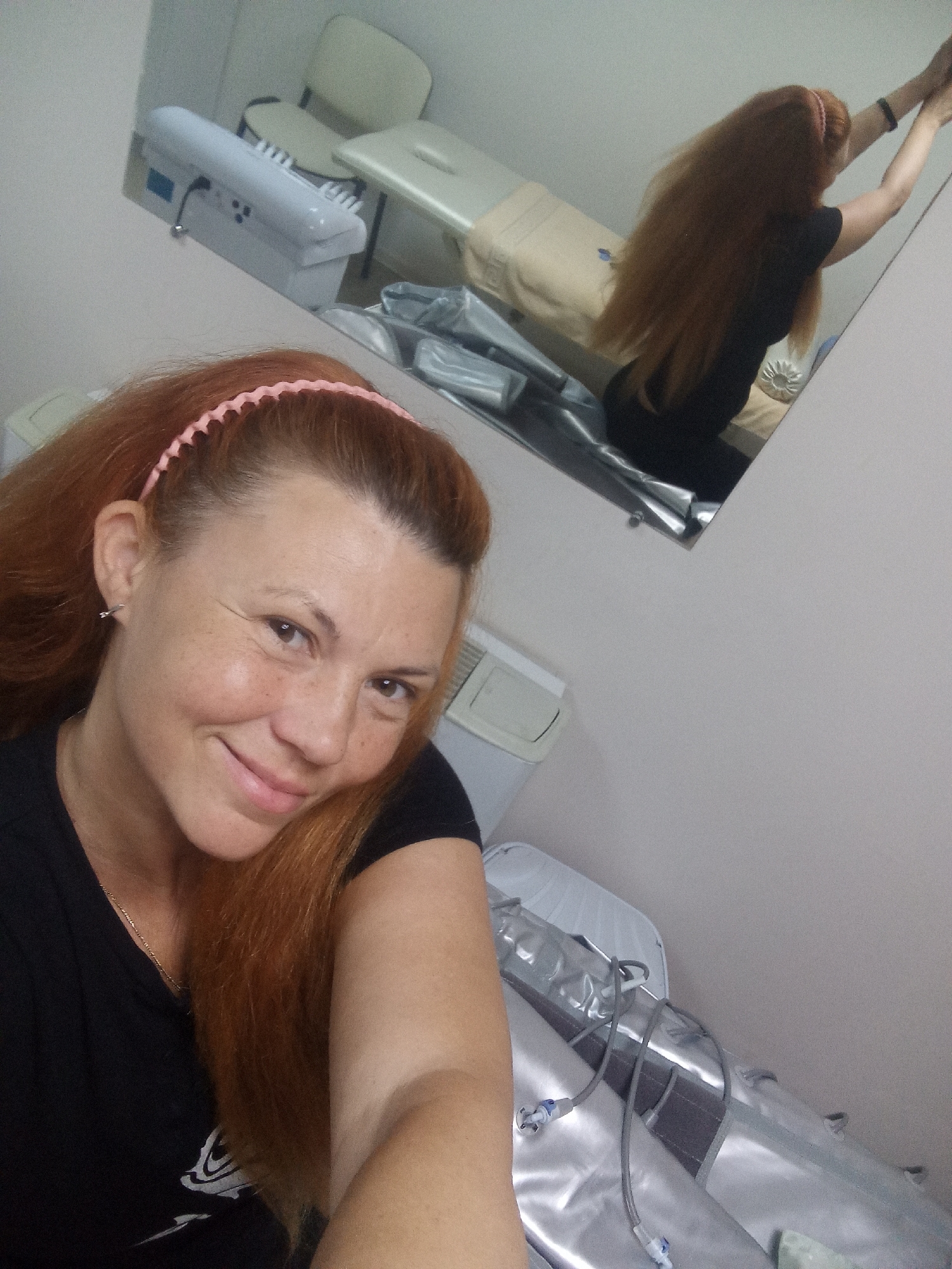 A woman with long, auburn hair and a pink headband is taking a selfie in a room with a mirror. She is wearing a black t-shirt and a gold chain necklace. In the mirror, her reflection shows her in a black t-shirt and black pants, reaching her hands up towards the mirror. The room appears to be a spa or treatment room, with a massage table, a white medical-looking device, and a silver, inflatable-looking device with tubes connected to it. The lighting is neutral, and the overall scene is a casual snapshot.