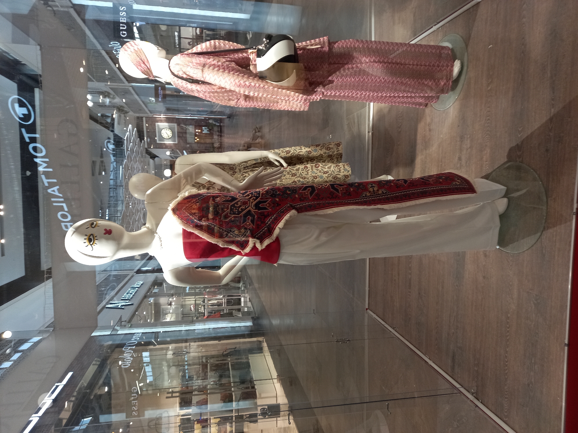 The image displays a retail store window with mannequins showcasing clothing. Three mannequins are visible, each wearing different outfits. The topmost mannequin is dressed in a pink patterned top and trousers. Below it, another mannequin wears a patterned skirt and a white top. The lowest mannequin is adorned in white trousers, a red top, and a decorative, possibly embroidered, vest with a red and black pattern. The mannequins are positioned behind a glass window, with reflections of the interior of the store and the exterior environment visible. Behind the mannequins, glimpses of store displays, shelves, and lighting are present. The floor of the display area appears to be made of wood-effect laminate. Visible text includes the brand name "GUESS" reflected on the glass, and partially visible store signage that reads "FABRIC MO" and "TOMOT". The scene suggests a fashion display, likely in a mall or shopping street, given the presence of multiple storefronts in the reflection. The lighting suggests it is daytime.
