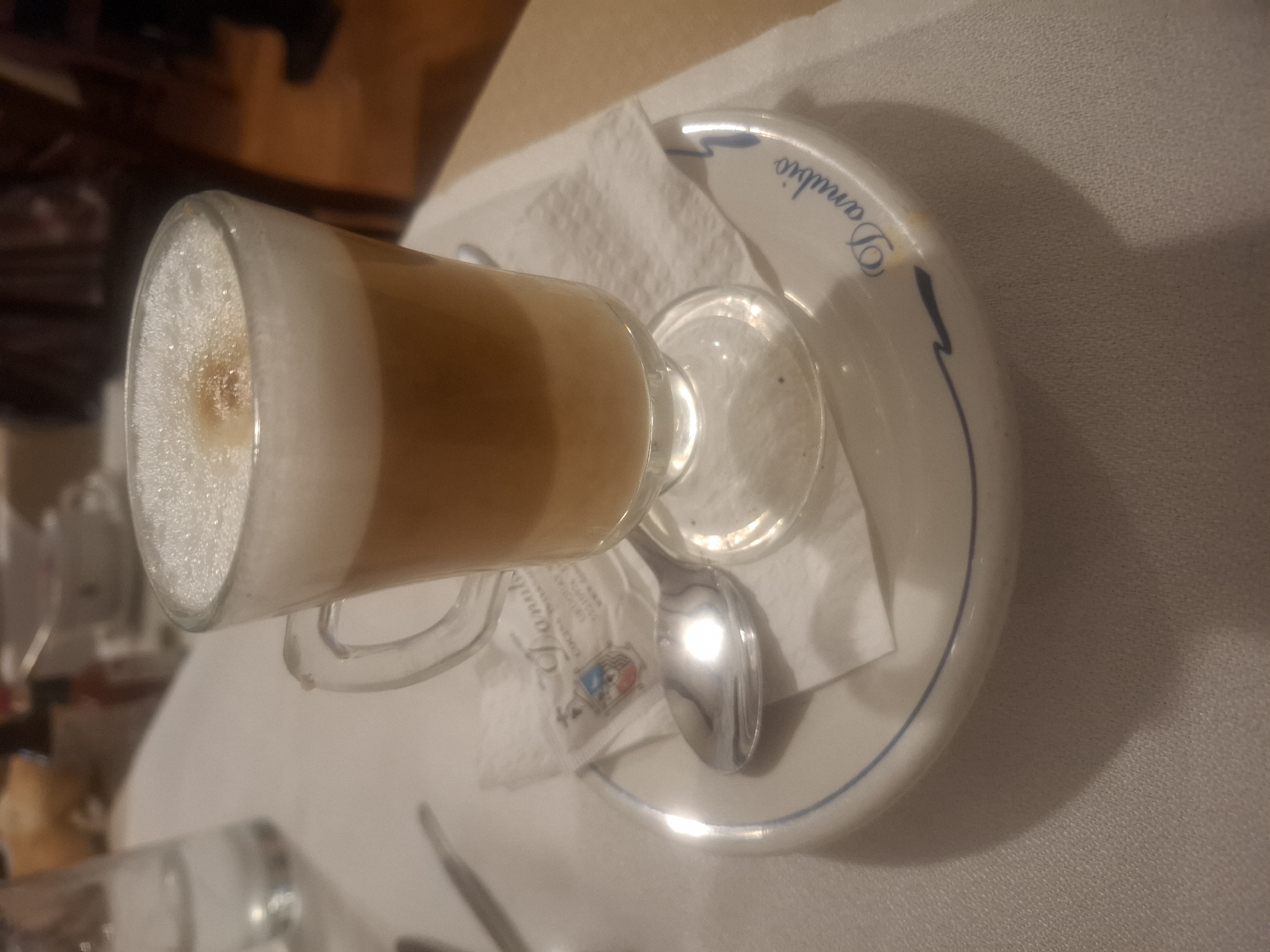 A tall glass of coffee with a foamy top sits on a white plate with the inscription "Danubio" in blue lettering. The plate rests on a white napkin, alongside a small sugar packet and a silver spoon. The scene is situated indoors, likely in a café or restaurant. The location is identified as Mexico City, Mexico, based on the inscription "Danubio", a well-known café in the city. No people are visible in the immediate view. Other objects, such as a glass bottle and other items, are partially visible in the background, suggesting a table setting. The lighting is somewhat dim.