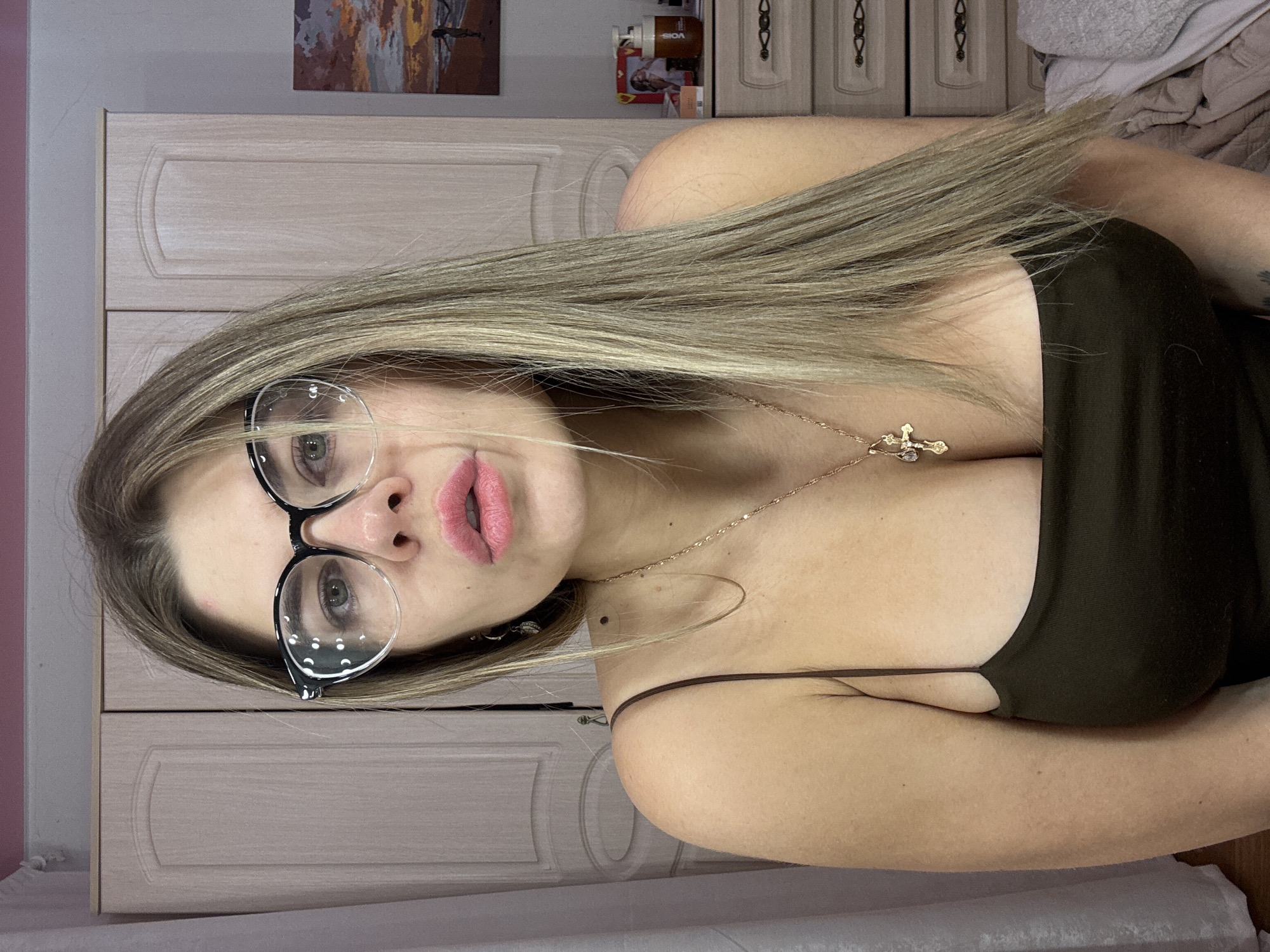 The image depicts an adult female subject positioned centrally, directly facing the camera in an indoor setting. She has long, straight, blonde hair, light-colored eyes, and fair skin. She wears black-framed glasses with clear lenses and a dark green or olive-colored spaghetti-strap top, displaying cleavage. A gold-colored chain necklace with a crucifixion crucifix pendant is worn around her neck. Her lips are pursed, covered with pink lipstick. A small, reddish mark is visible on her forehead. On her left ear, a small, metallic earring is partially visible. The specific location within the city of Chaikovsky, Russia, is not discernible from the visual evidence present in the image.
The background consists of light-colored wooden furniture. To the subject's left is a two-door wardrobe or cabinet with decorative paneling. To her right is a multi-drawer dresser or nightstand with dark, decorative handles. On top of this dresser, a small framed picture depicting a sunset over a beach with figures is visible, alongside a pump bottle labeled "VOIS" and other indistinct items. The wall behind the subject is light grey or beige, with a horizontal strip of pink paint visible near the ceiling. A light-colored curtain is visible on the far left, and a crumpled light grey blanket or fabric is visible in the bottom right corner, suggesting a bed. Lighting is even and diffused.