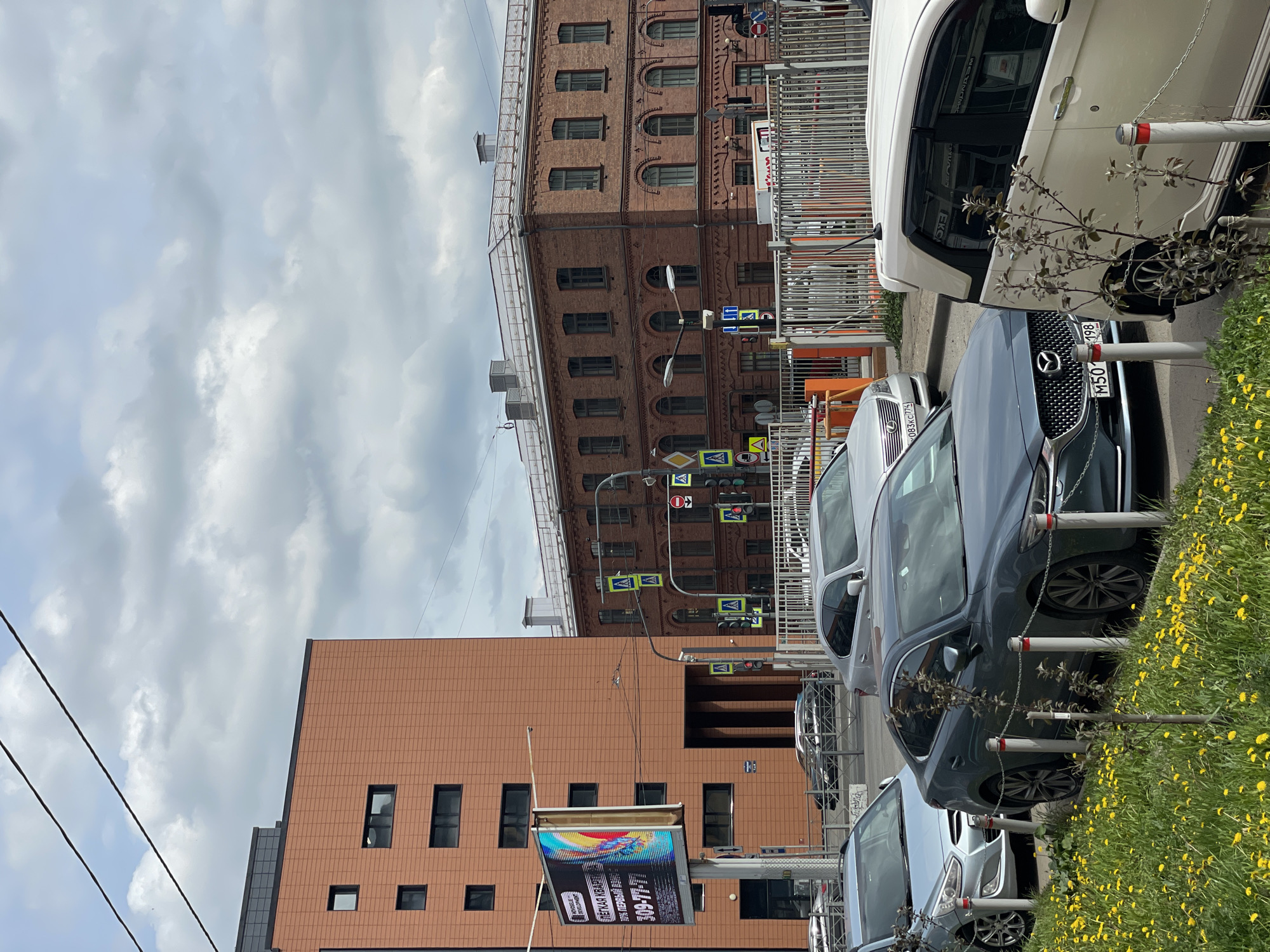 The image depicts a street scene in Saint Petersburg, Russia, characterized by buildings of varying architectural styles and parked cars lining the road. The foreground is dominated by several cars parked along the roadside, adjacent to a small patch of grass interspersed with yellow flowers. Behind the cars, two prominent buildings are visible. One is a modern structure with a tiled facade and rectangular windows. The other is an older, taller brick building with arched windows and classical architectural details. Street signs and traffic lights are suspended above the road. The sky is overcast with heavy cloud cover. A large advertising billboard is present near the modern building.