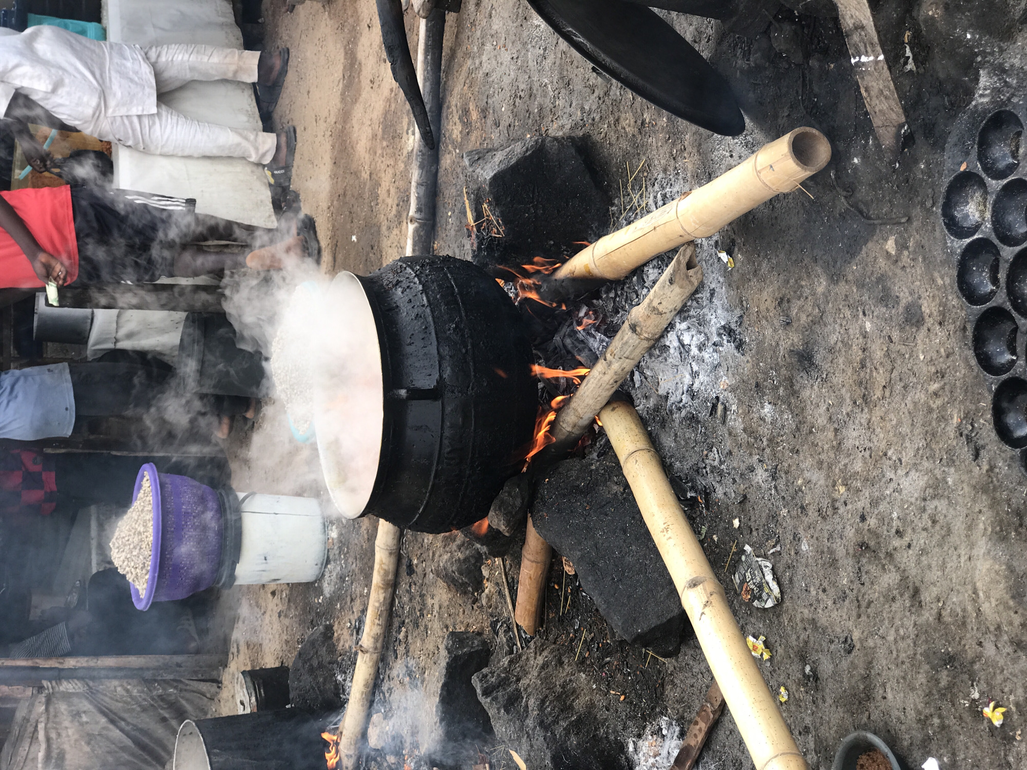 In Dutsen Alhaji, Nigeria, a large, black, cauldron-like pot sits atop an open fire supported by bamboo poles and rocks. The pot emits copious amounts of steam. Nearby, a purple plastic basket filled with a light-colored, granular substance rests atop a white bucket. Several people are standing in the background, their bodies partially obscured by the steam. To the right, a black pan with multiple circular indentations lies on the ground. The ground is dirt and ash.
