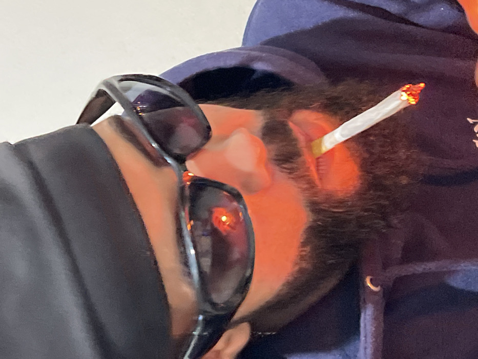 A close-up, chest-up image shows an individual, appearing male, with a full, dark beard. The person wears a black bandana or head covering and dark, wraparound sunglasses. A dark, likely navy blue, hooded sweatshirt is visible, with a drawstring eyelet on the left side of the collar. A hand-rolled cigarette, actively burning with a red-orange ember, is held between the lips. The lower face, beard, and the lit cigarette are illuminated by a strong, warm orange light, which is also distinctly reflected as a bright red-orange spot in both lenses of the sunglasses. The background is indistinct and light-colored. The specific location within the city of Tanger, Morocco, cannot be determined from the visual information provided in the image.