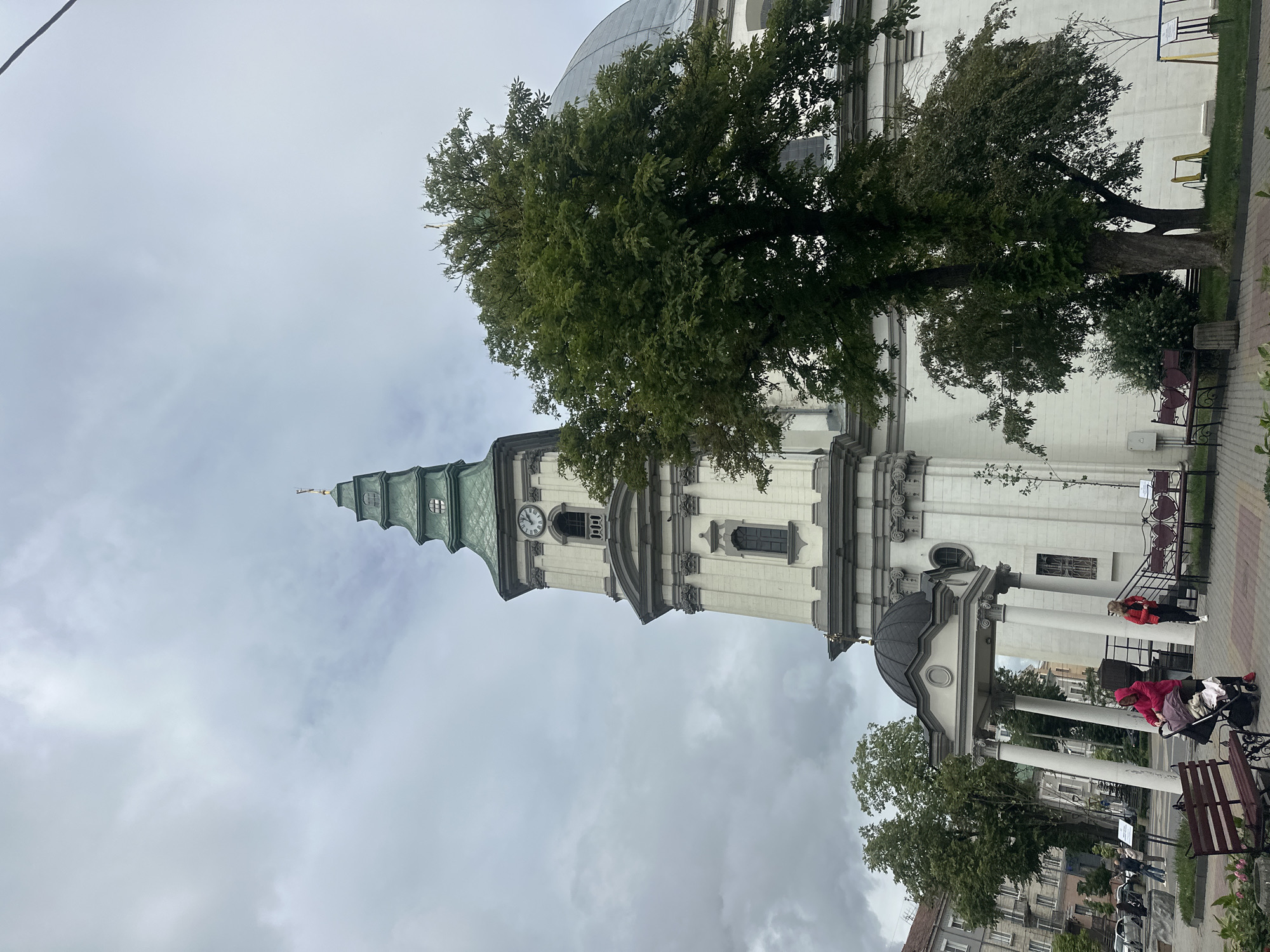 The image depicts the Ternopil Cathedral of the Immaculate Conception of the Blessed Virgin Mary in Ternopil, Ukraine. The grand white facade of the cathedral dominates the right side of the frame, with a tall, slender tower topped by a green spire on the left. A large, leafy green tree is positioned in front of the cathedral, partially obscuring its dome. The sky is overcast with heavy, gray clouds. In the foreground, a paved area features benches, and several people are visible. Two women are the most prominent: one in a pink hooded jacket is pushing a stroller, and another in a red jacket stands near a set of stairs. Cars are parked in the background, and the surrounding urban environment is suggested by buildings visible to the left. The overall scene conveys a sense of an overcast day in a European city.
