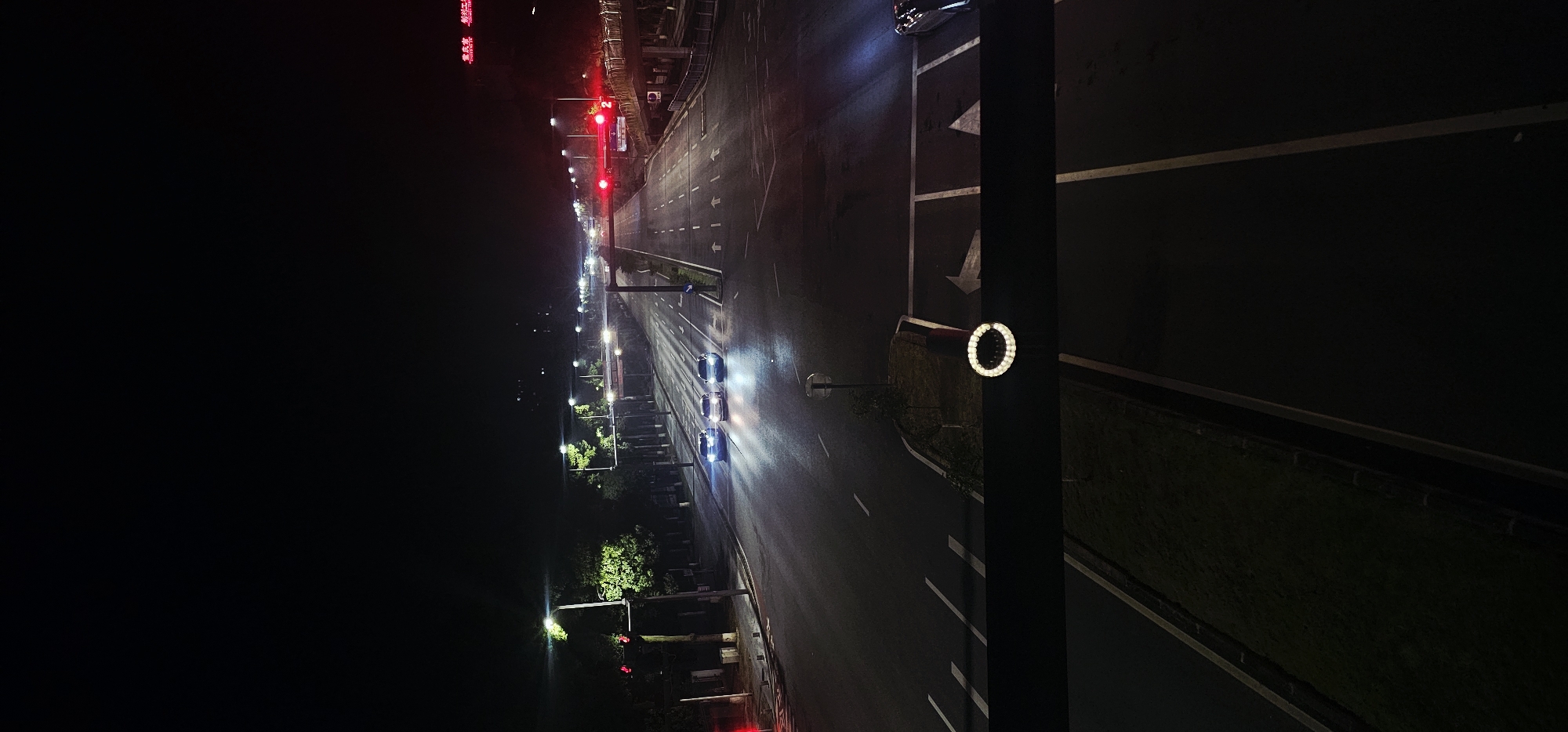 The image shows a nighttime view of a multi-lane road in the Chongqing High-tech Zone, China. Several cars are driving on the road, their headlights illuminating the asphalt. Streetlights and traffic lights cast a glow on the scene, creating reflections on the wet road surface. Trees line the side of the road, and a pedestrian overpass is visible in the distance. A circular illuminated sign is affixed to a dark horizontal beam in the foreground. The overall scene depicts a typical urban streetscape at night, with the characteristic lights and infrastructure of a modern city.