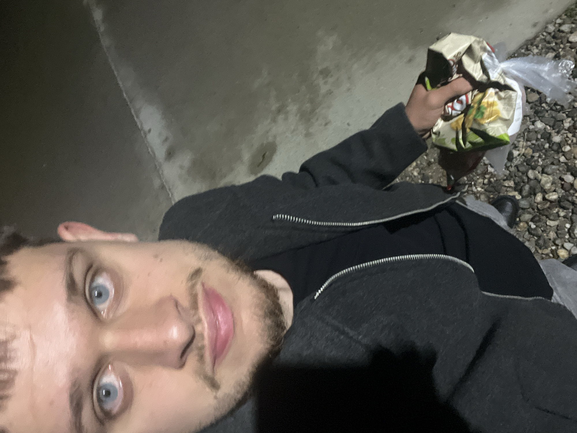 A person is holding a partially crushed bag of Doritos in their right hand. The person is wearing a black shirt and a gray jacket with a zipper. The person has light blue eyes and stubble on their face. The location appears to be outdoors, possibly near a sidewalk and an area covered with small rocks. The lighting is dim, suggesting it is nighttime. The location is not specified, and the claim that the image was taken in the city of Blagoevgrad, Bulgaria, is unconfirmed.